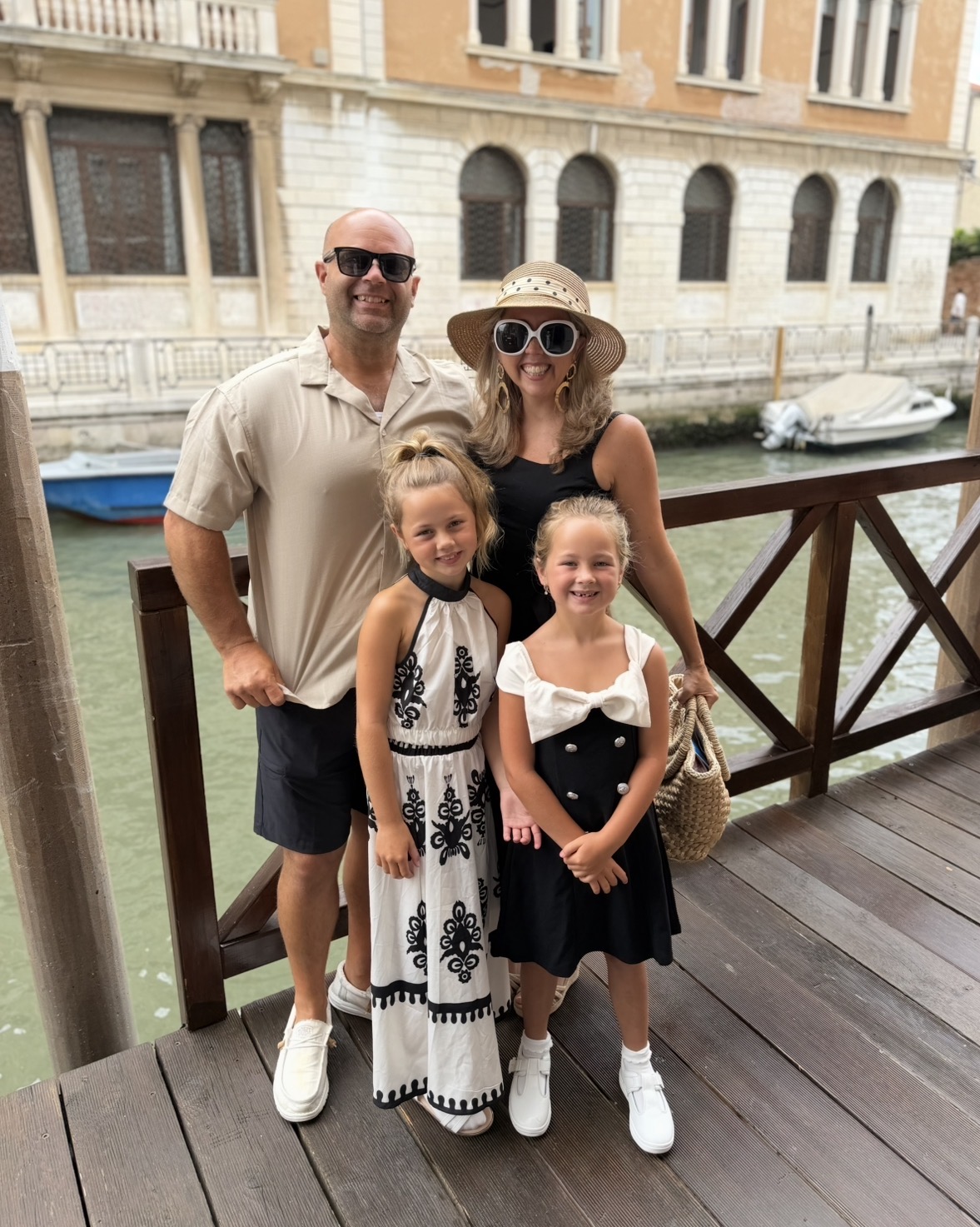 Family of four standing on a wooden bridge by a canal, smiling, with historic buildings in the background.