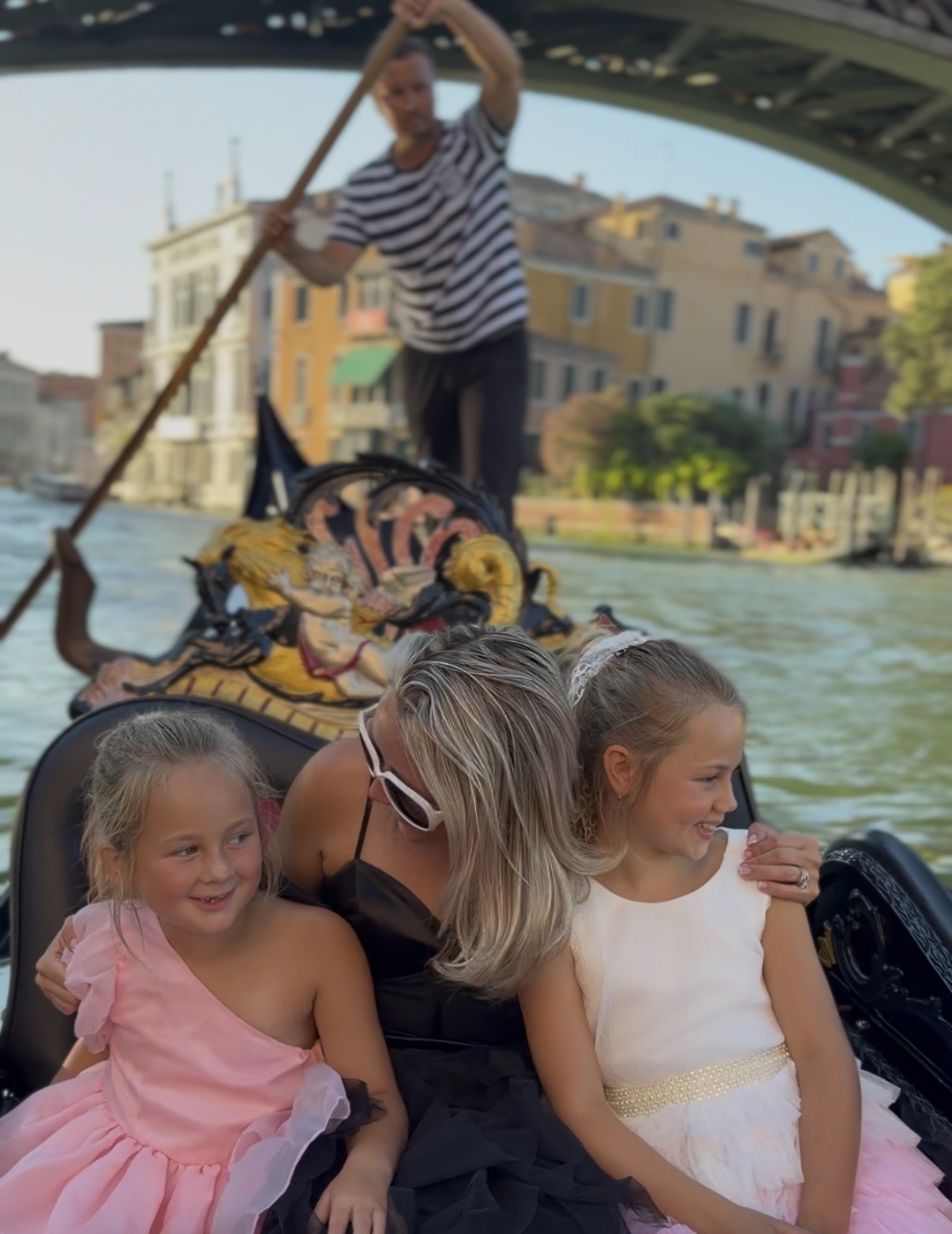 Two young girls sitting in a boat with a woman, a gondolier paddling in the background, and buildings along a canal.