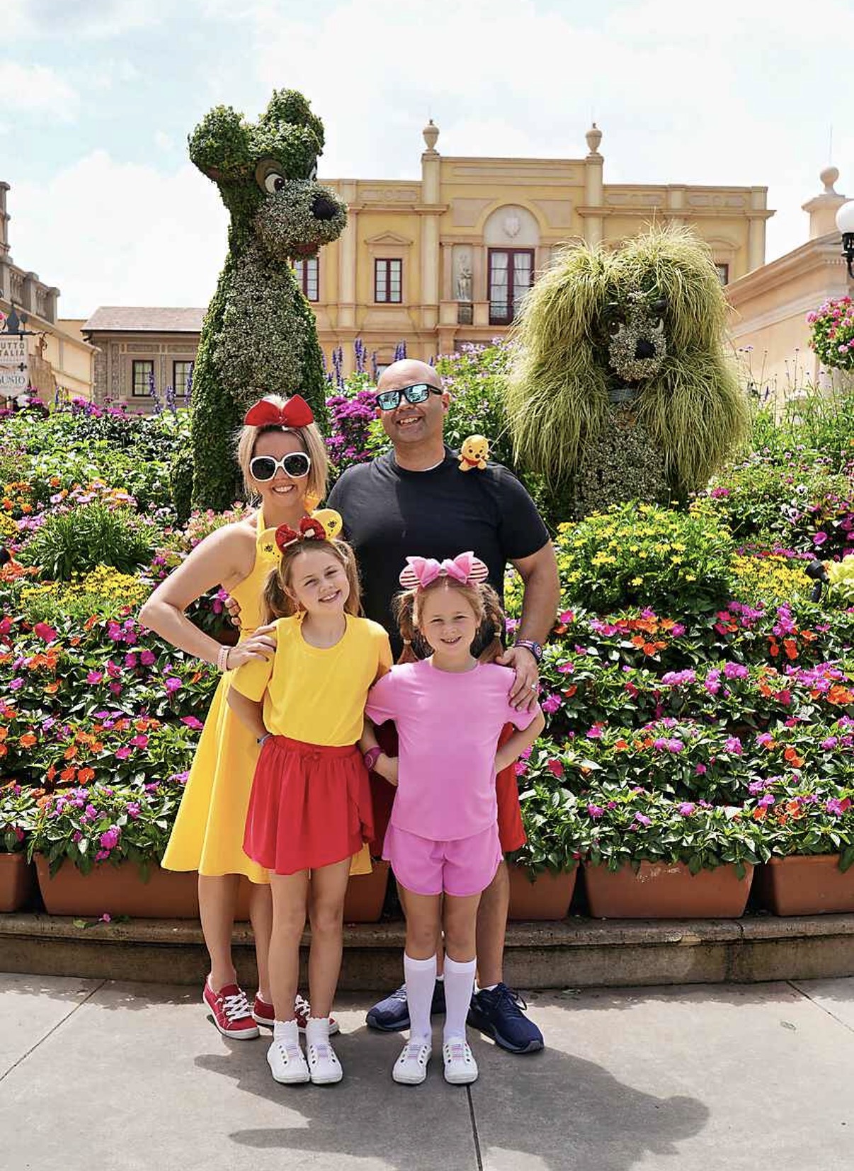 Group of two adults and two children standing in front of topiary animals and colorful flowers, with a building in the background.