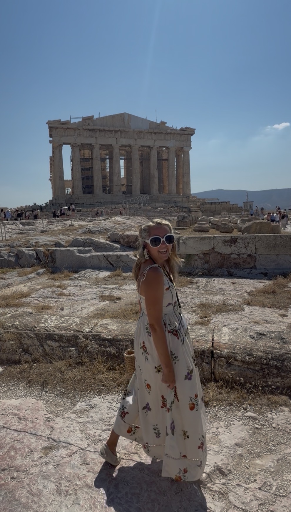 Woman in a floral dress and sunglasses standing in front of the Parthenon on a sunny day.