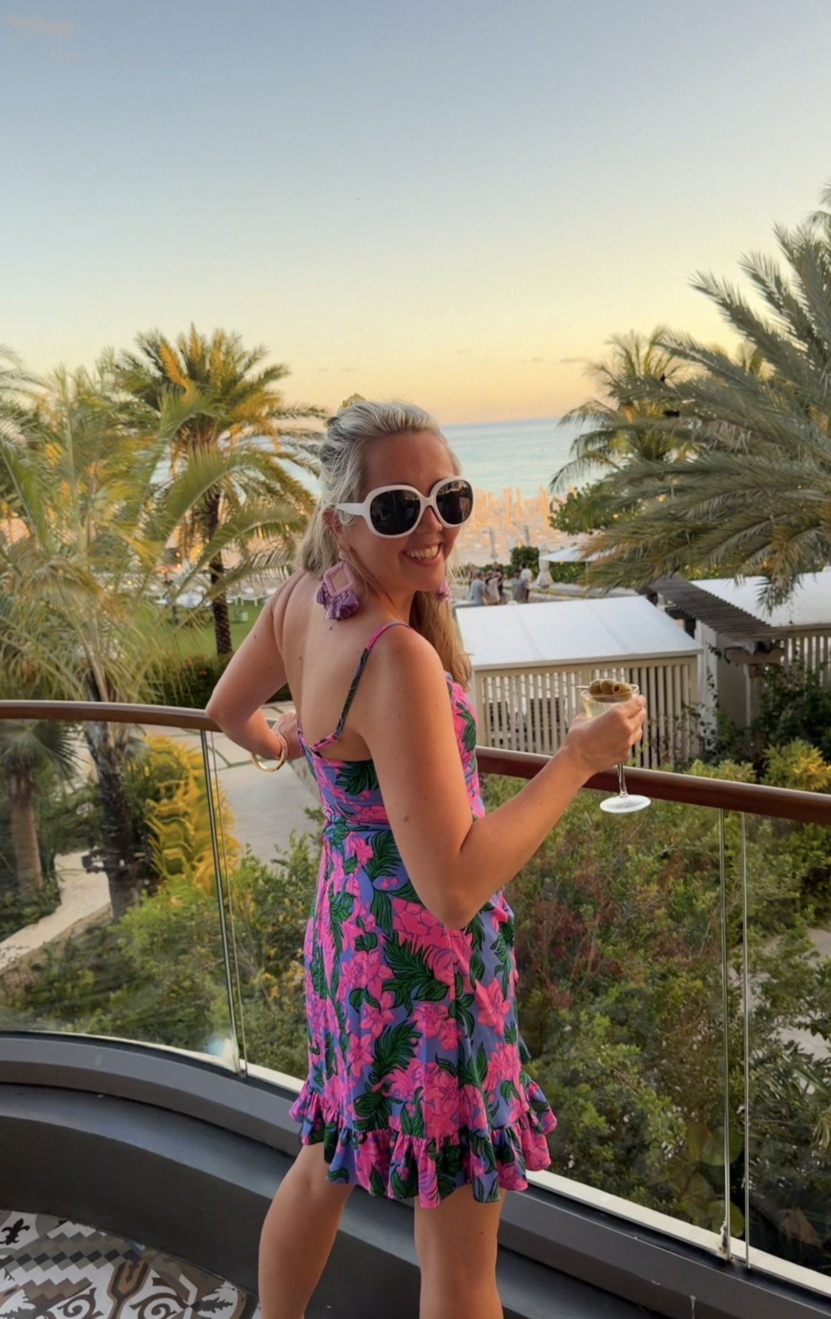 Woman in sunglasses and a colorful dress holding a drink on a balcony with palm trees and a sunset view.