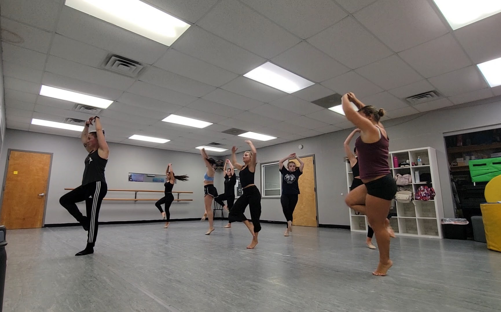 Group of people practicing dance or exercise in a studio with bright ceiling lights and shelves in the background.