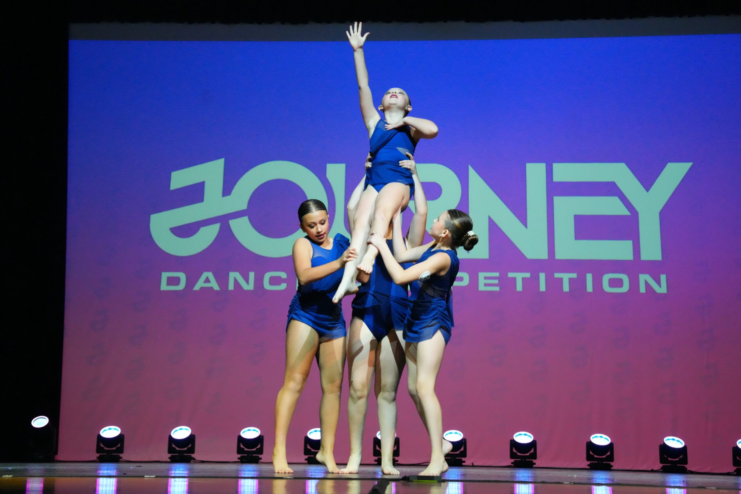 Four dancers in blue costumes perform a lift on stage with a pink and purple background and the word 'JOURNEY' in large letters.