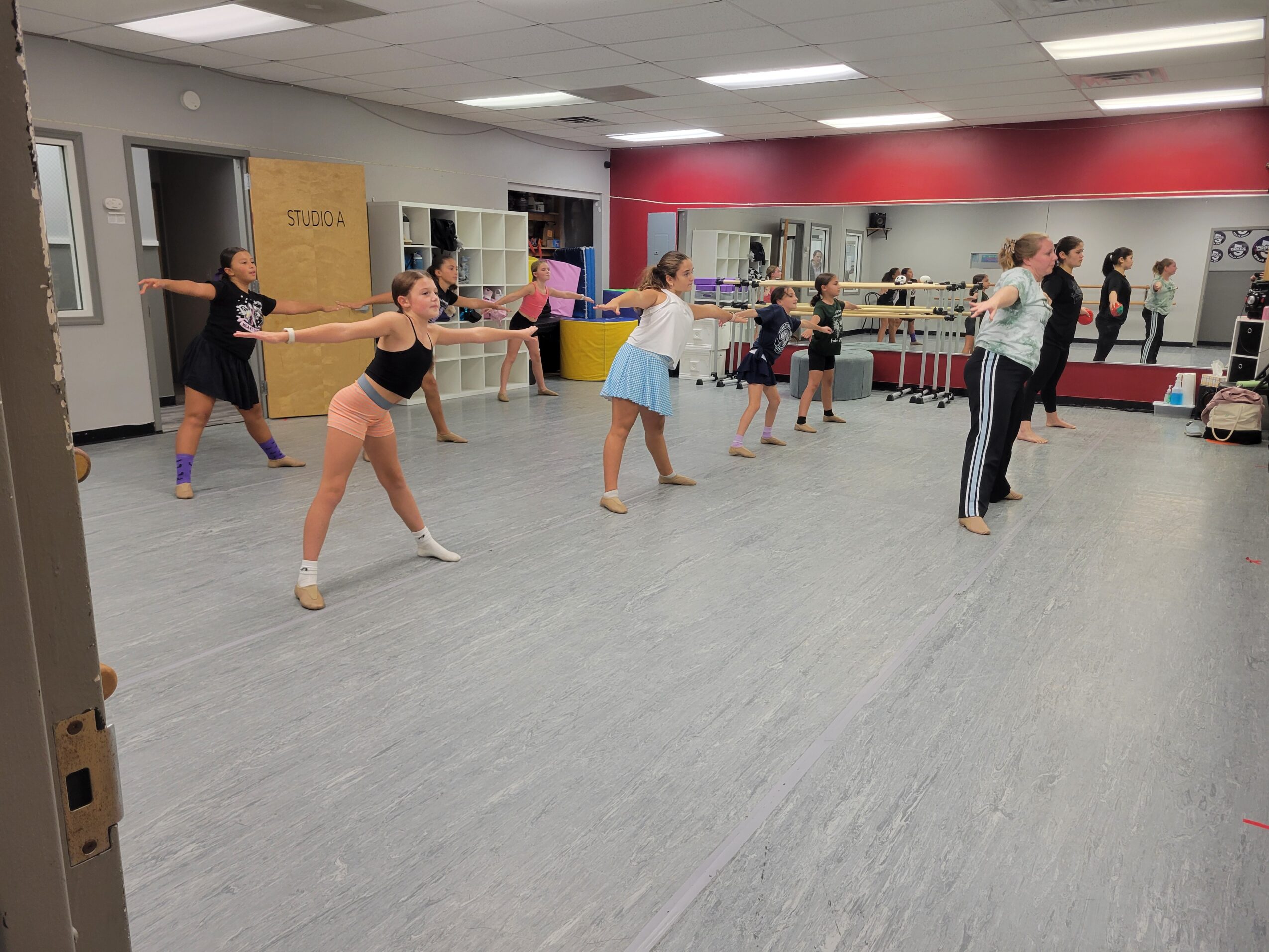 Group of children and an instructor in a dance studio practicing with a long stick, mirrors on the wall, and a red accent wall.