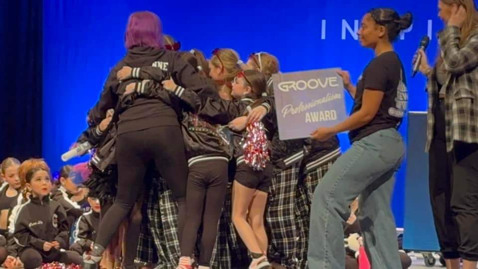 Group of children and adults on stage, some hugging, with a blue background and a sign reading 'GROOVE AWARD'.