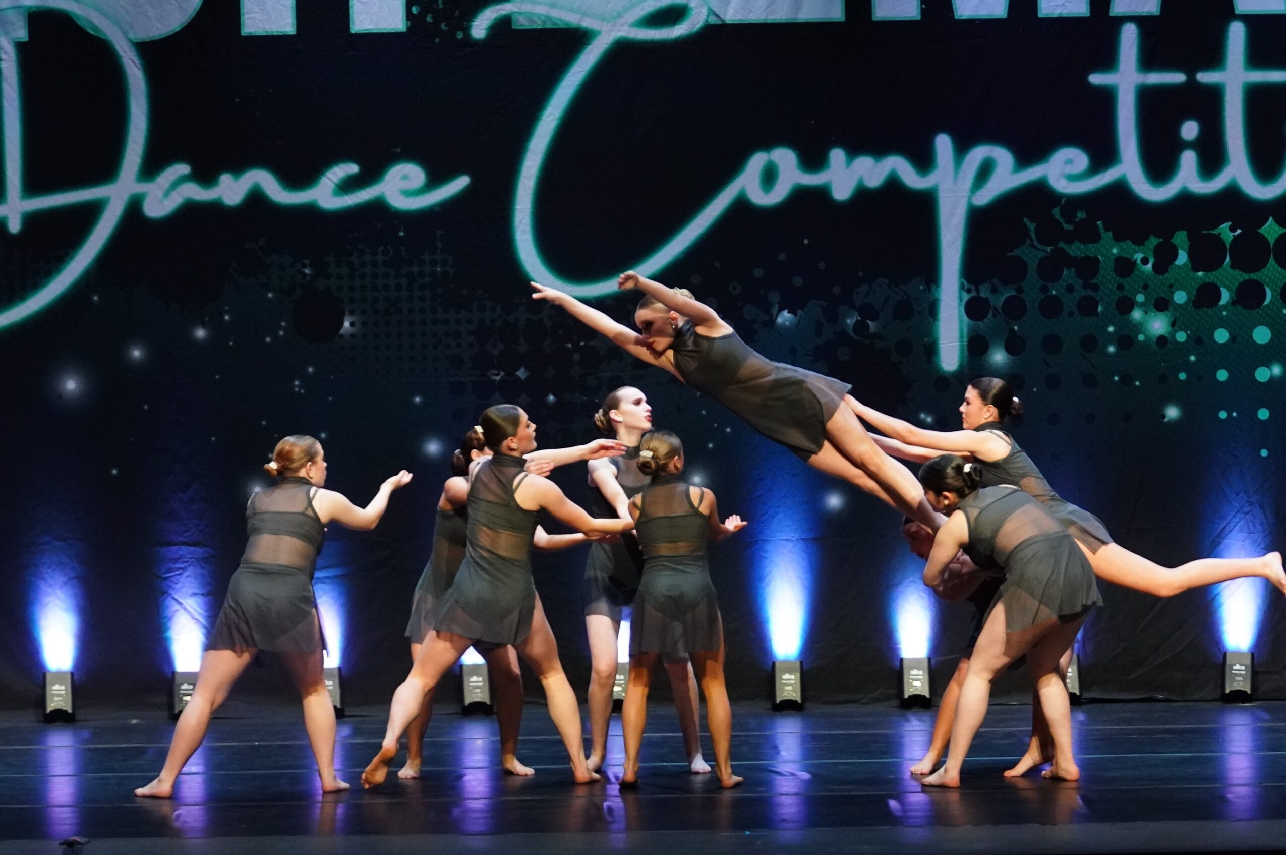 Group of dancers performing on stage with a dark background and blue lighting, some lifting others in a dance move.