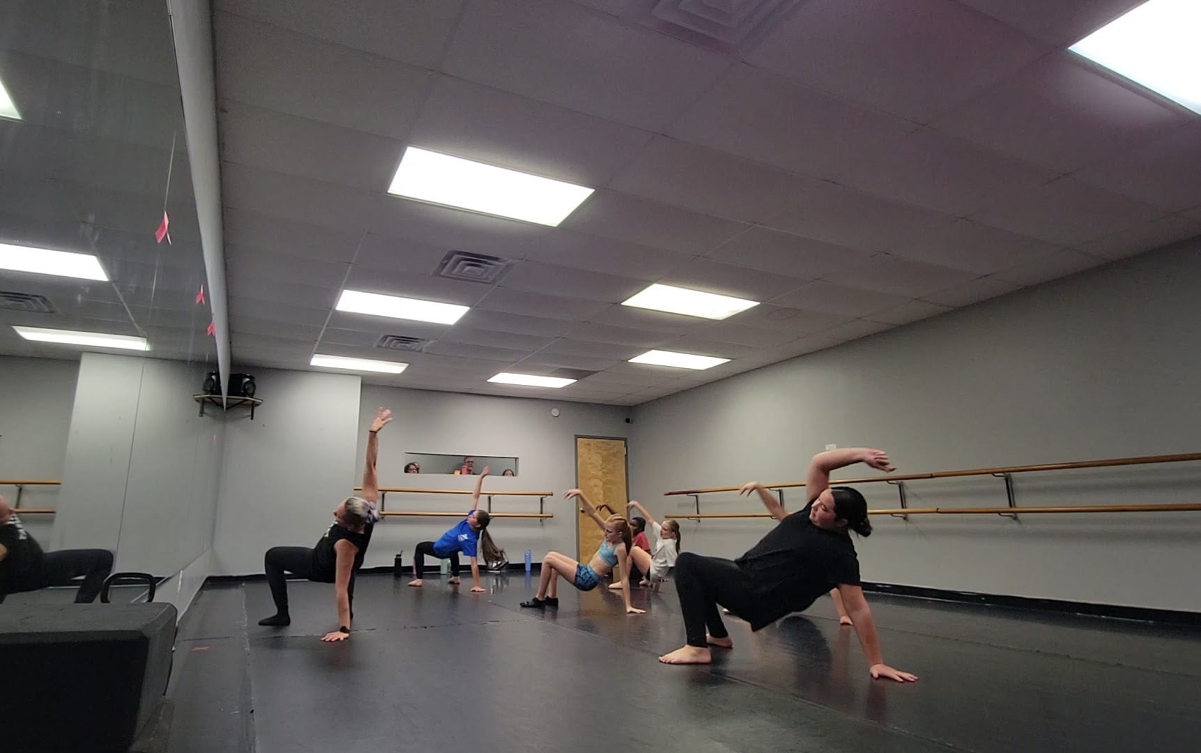 People stretching and exercising in a dance studio with mirrors and ballet barres, some on the floor and some standing.