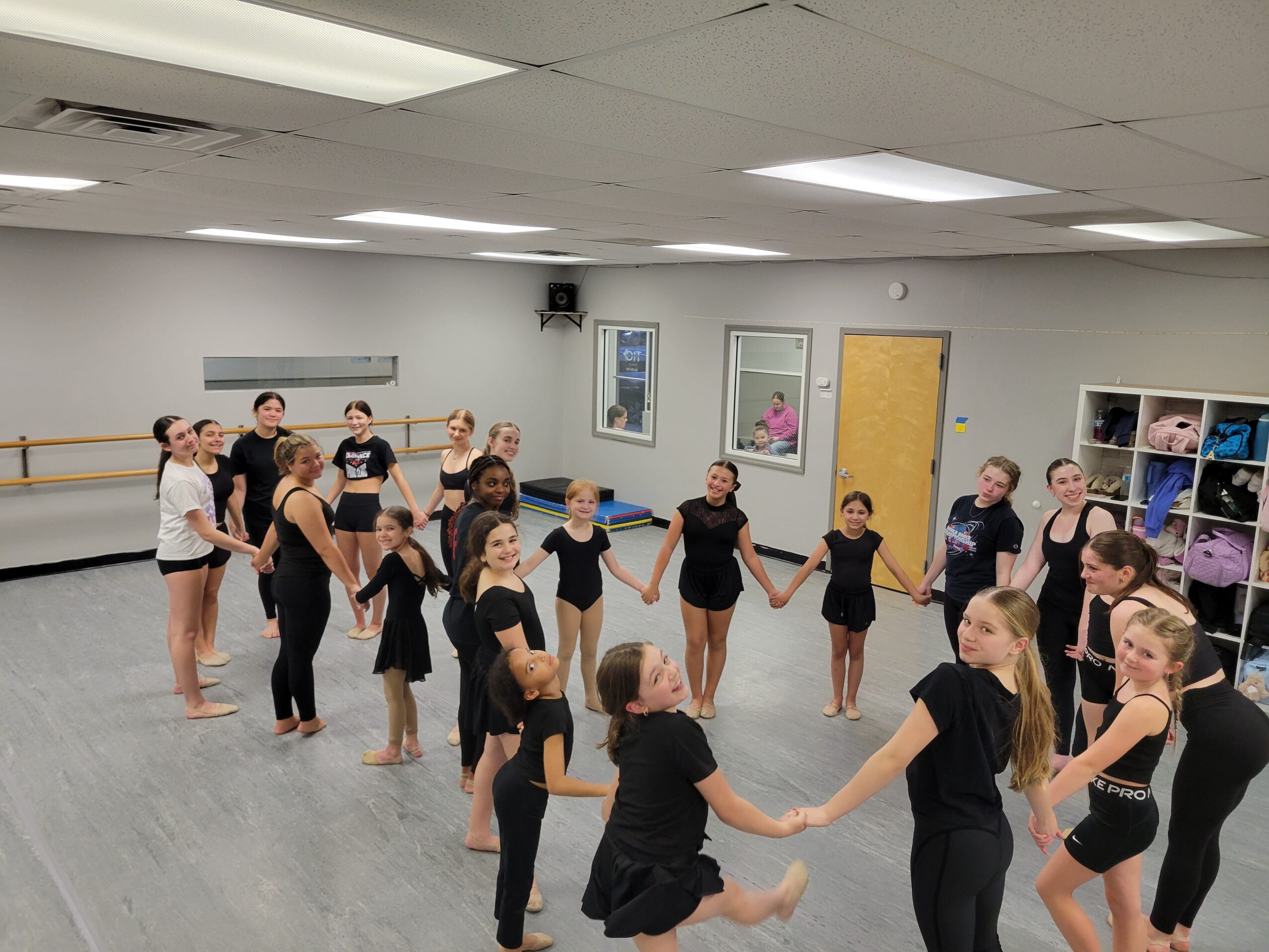 Group of young girls in black dance attire holding hands in a dance studio, practicing a routine.