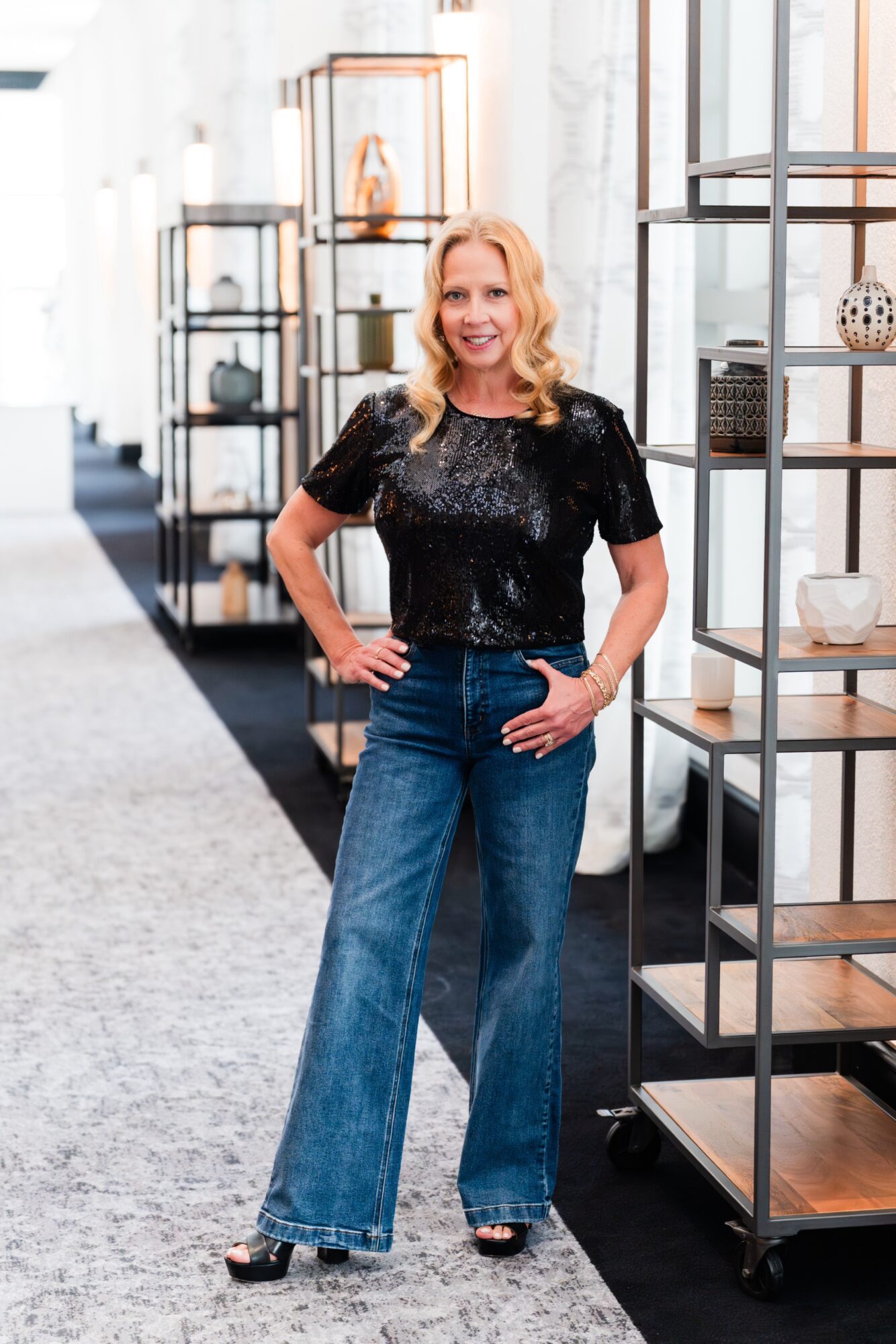 Woman with blonde hair wearing a black sequined top and wide-leg jeans standing in a modern store with shelves.