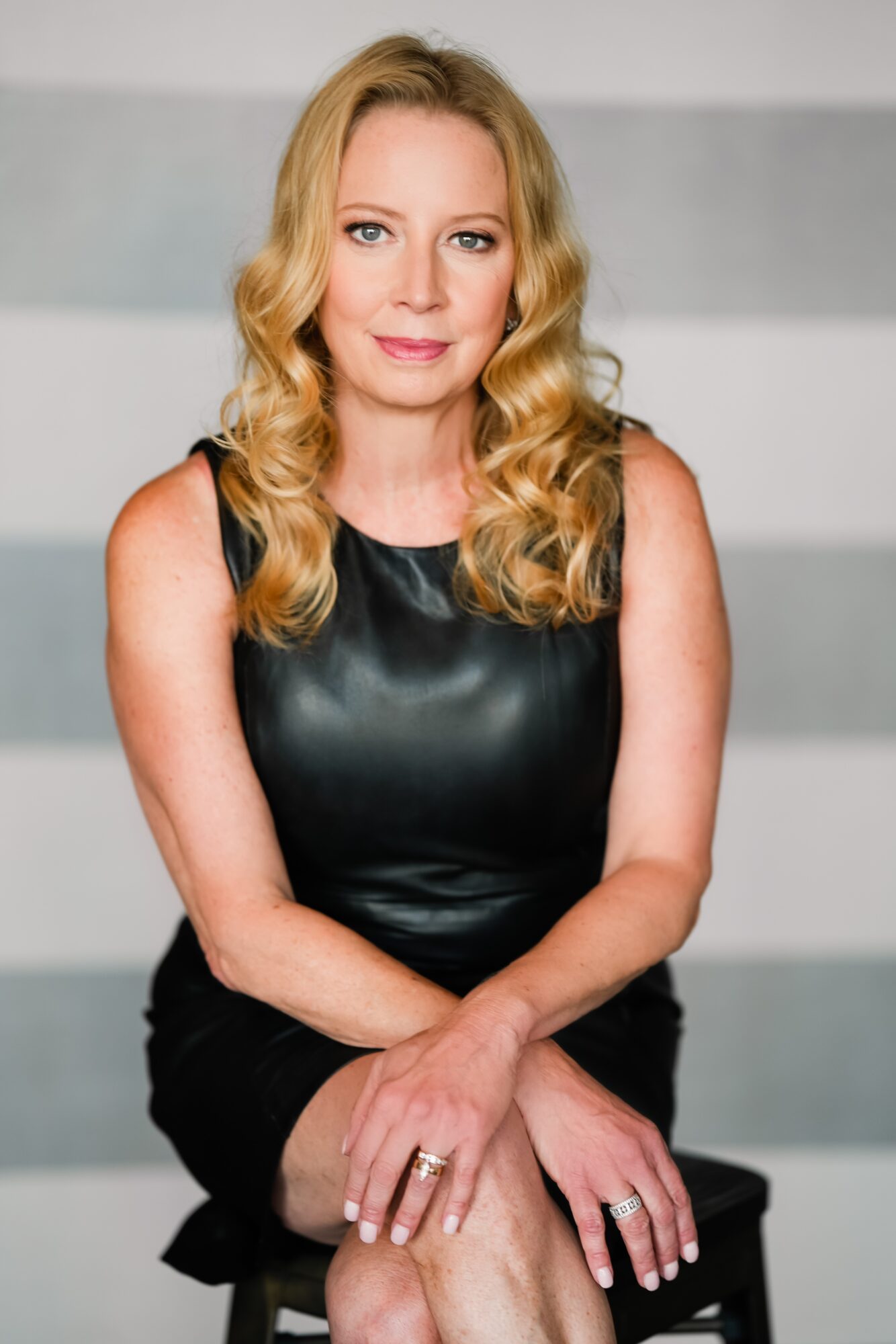 Woman with blonde hair in a black sleeveless top, sitting with hands on lap, neutral background.