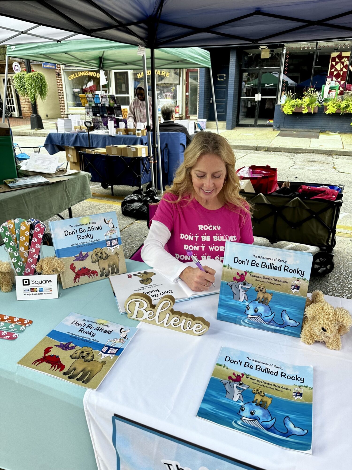 Woman with blonde hair sitting at a table under a tent, signing books about dogs and puppies, with books and decorations on the table.
