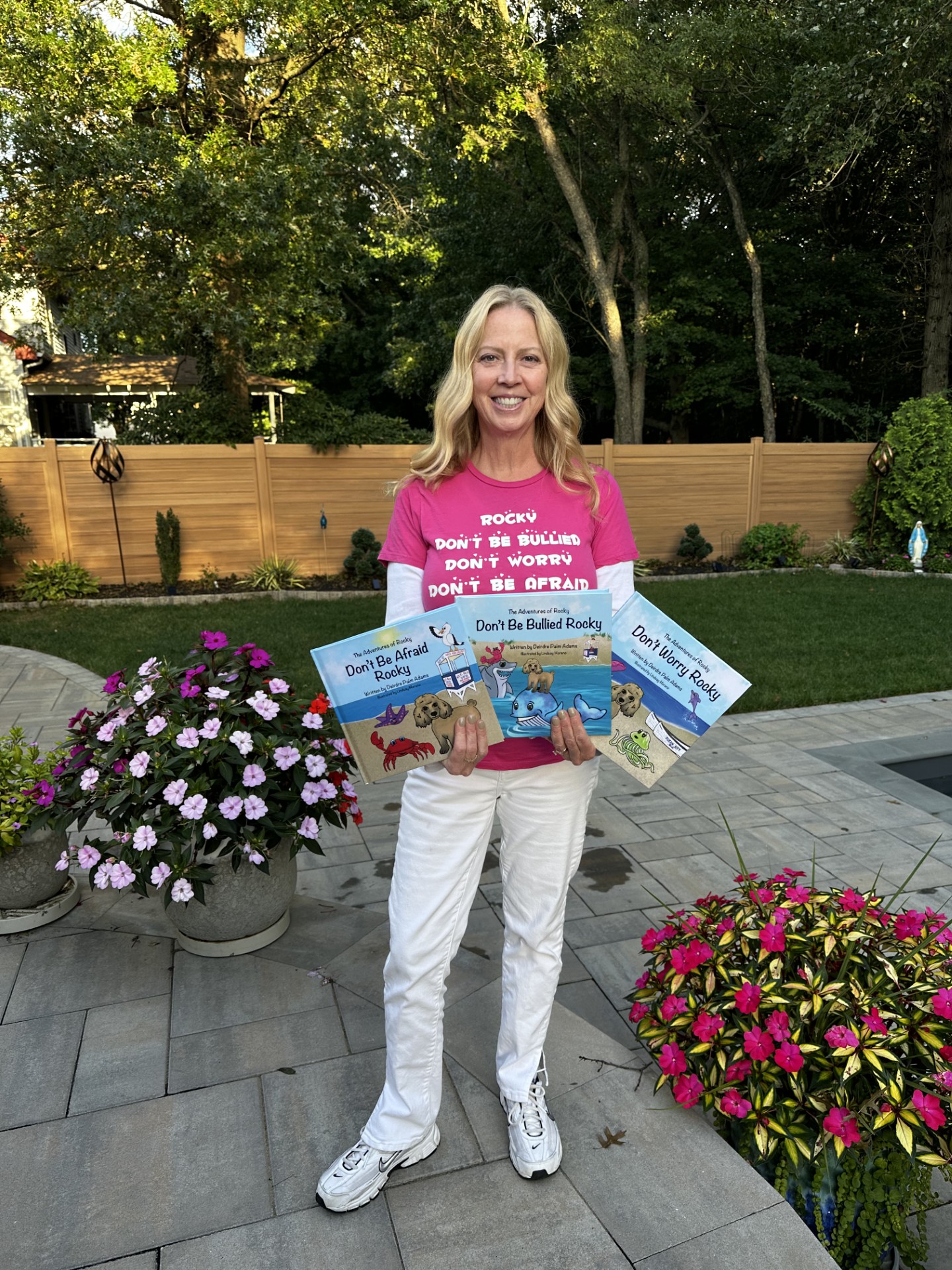 Woman standing outdoors holding books, smiling, with flower pots and trees in background.
