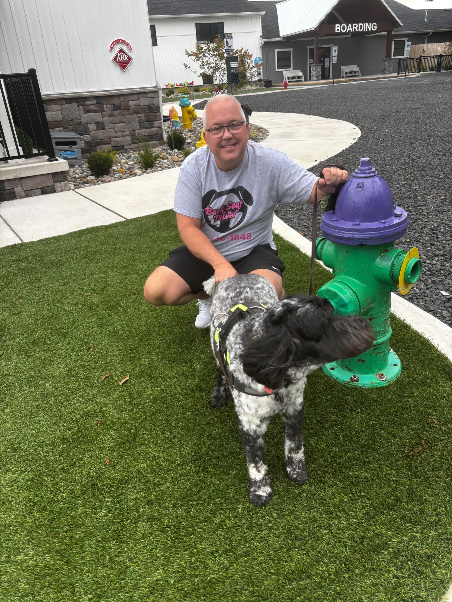 Person kneeling on grass with a black and white dog, colorful water play equipment nearby, urban background.