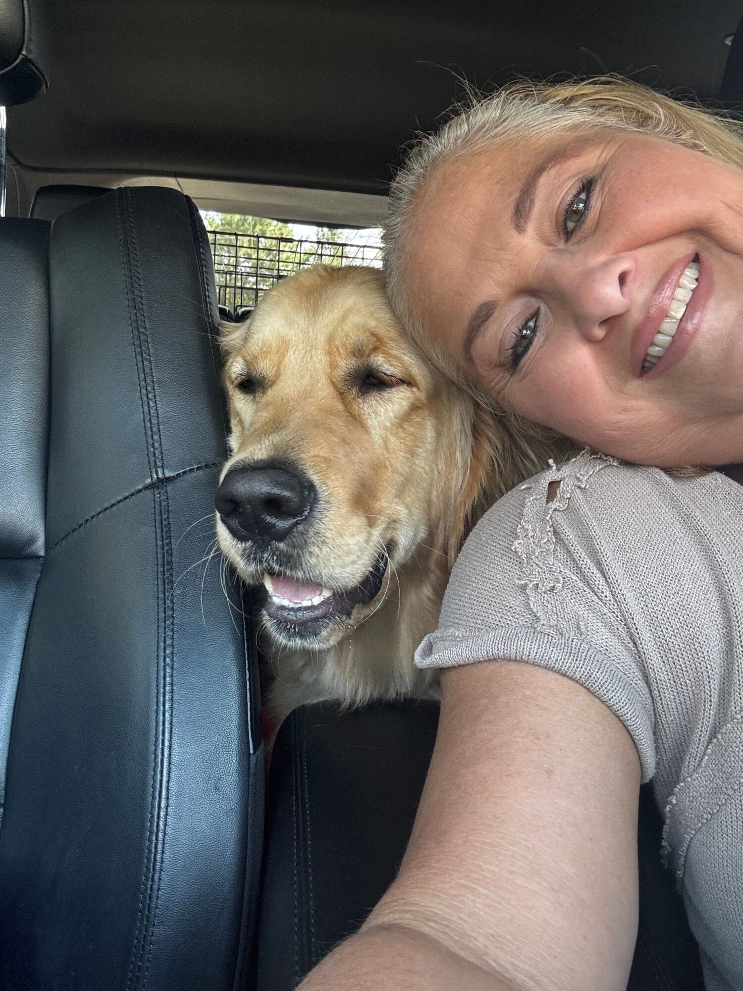 Person with blonde hair and a dog lying on a car seat, both smiling, in a close-up selfie.