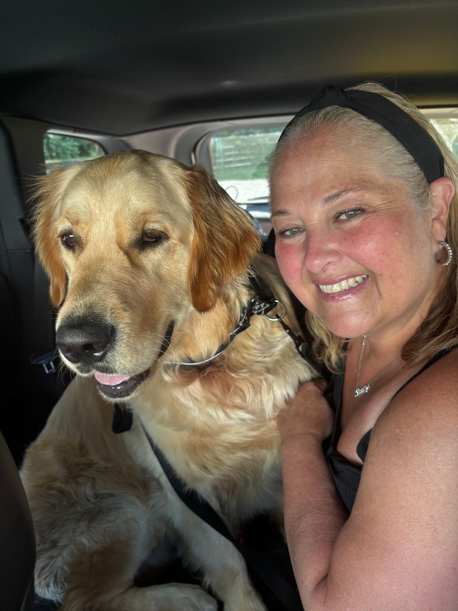 Woman with blonde hair and black headband smiling with a golden retriever dog inside a vehicle.