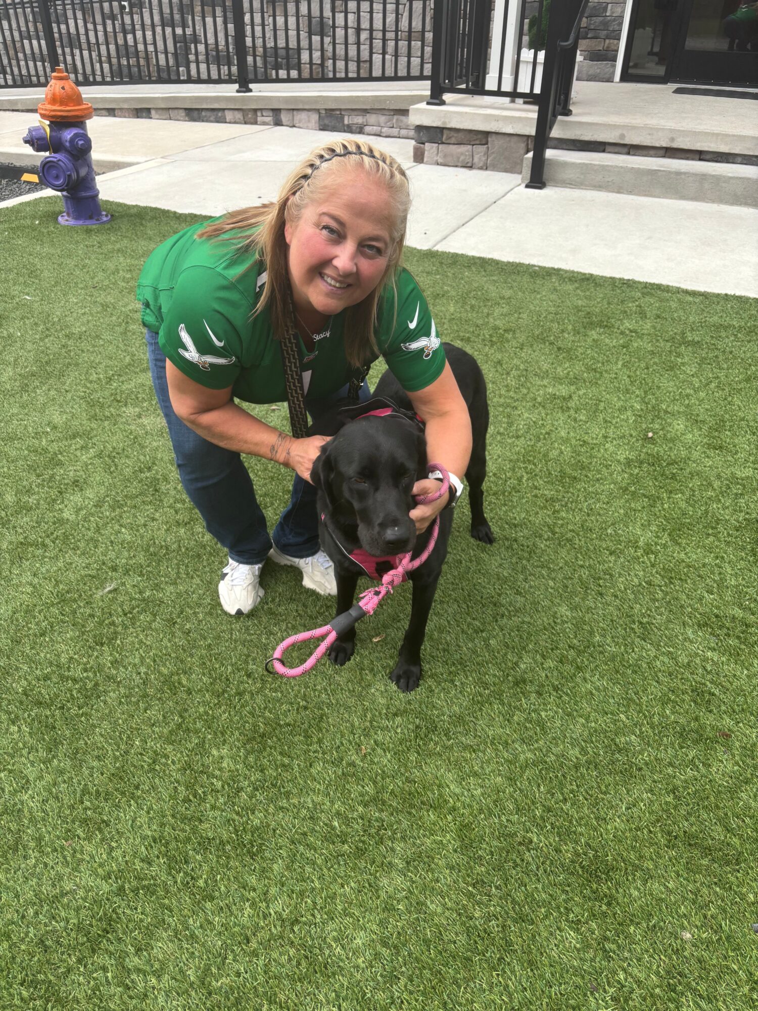 Woman crouching on grass with a black dog on a pink leash, near a sidewalk and fence.