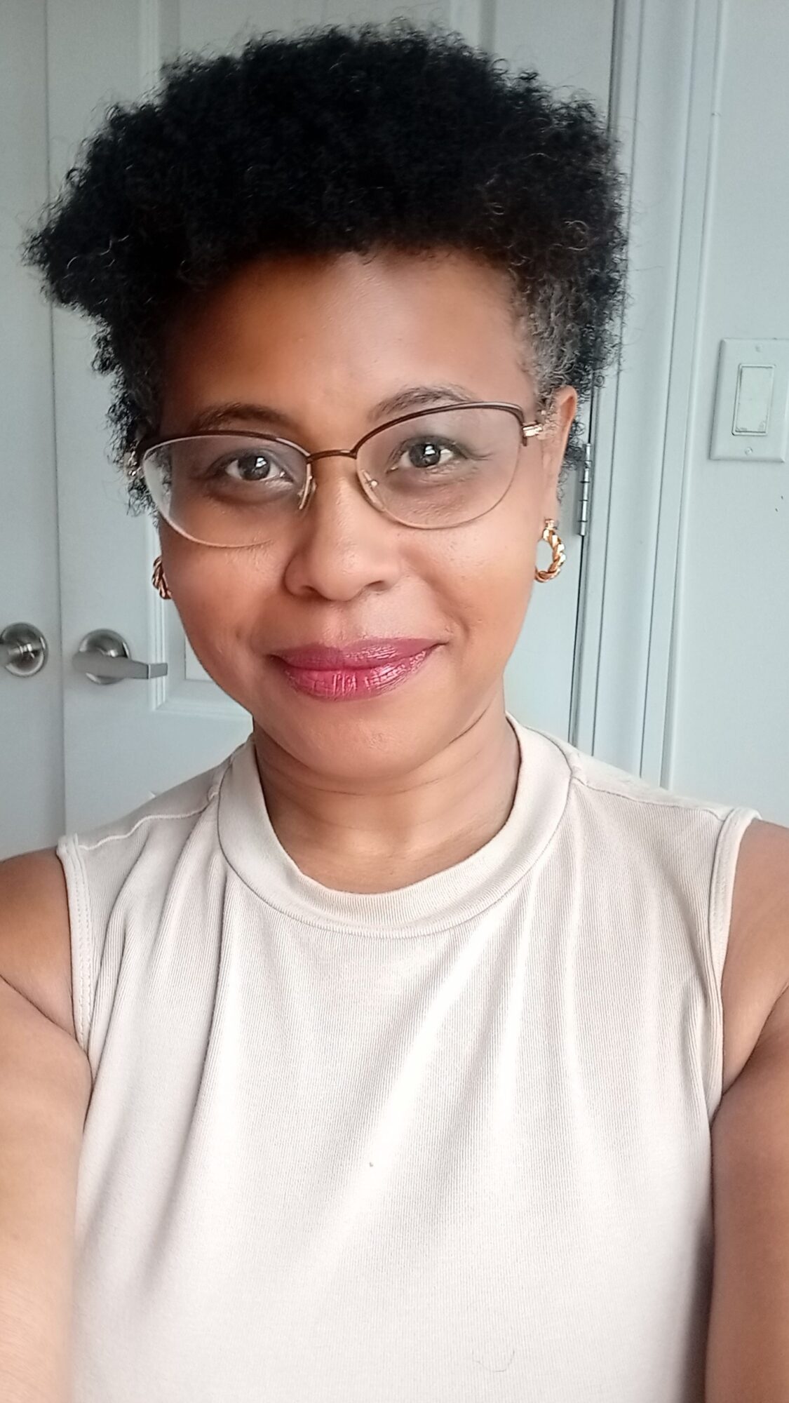 Portrait of a woman with glasses, short curly hair, wearing a sleeveless top, smiling, in front of a light-colored background.