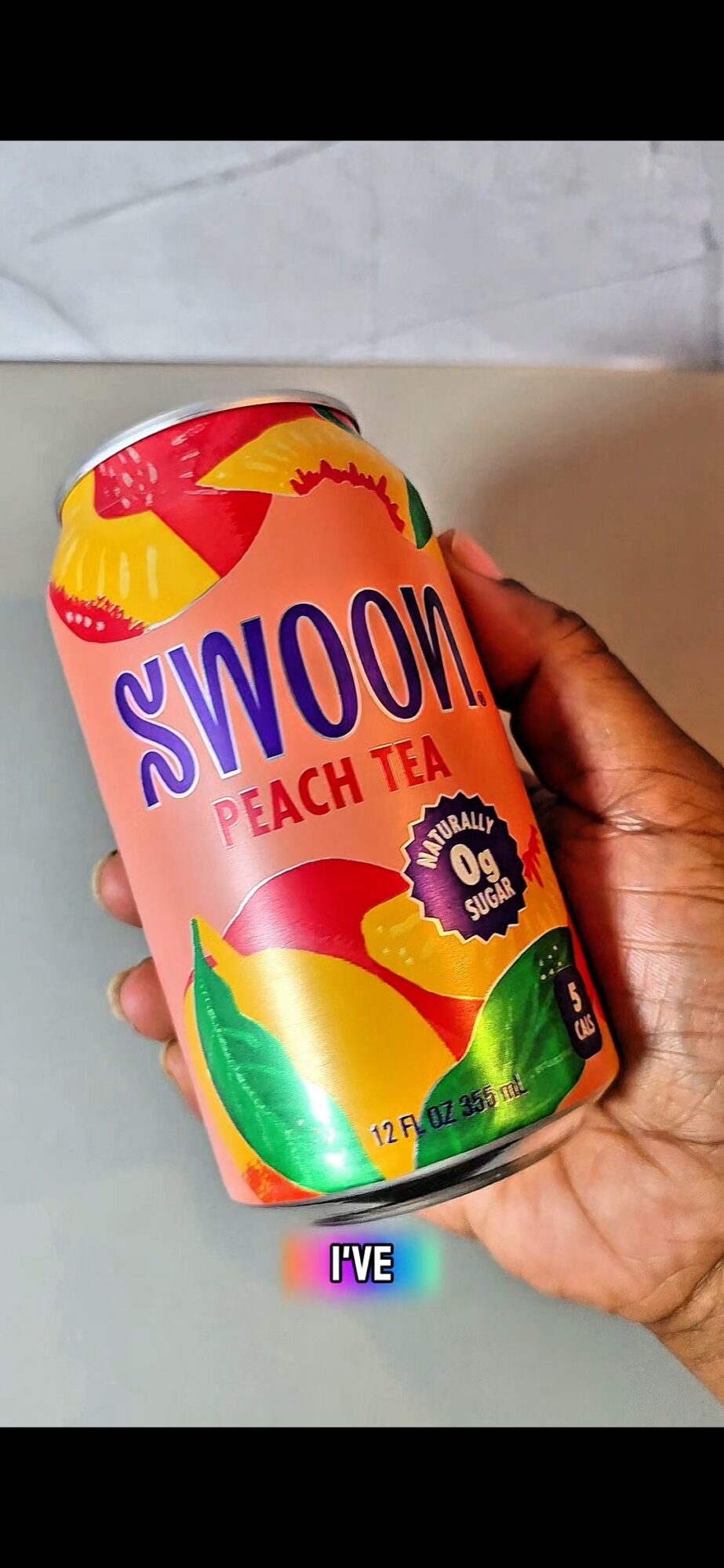 Hand holding a peach tea can with colorful design and text, against a light background.