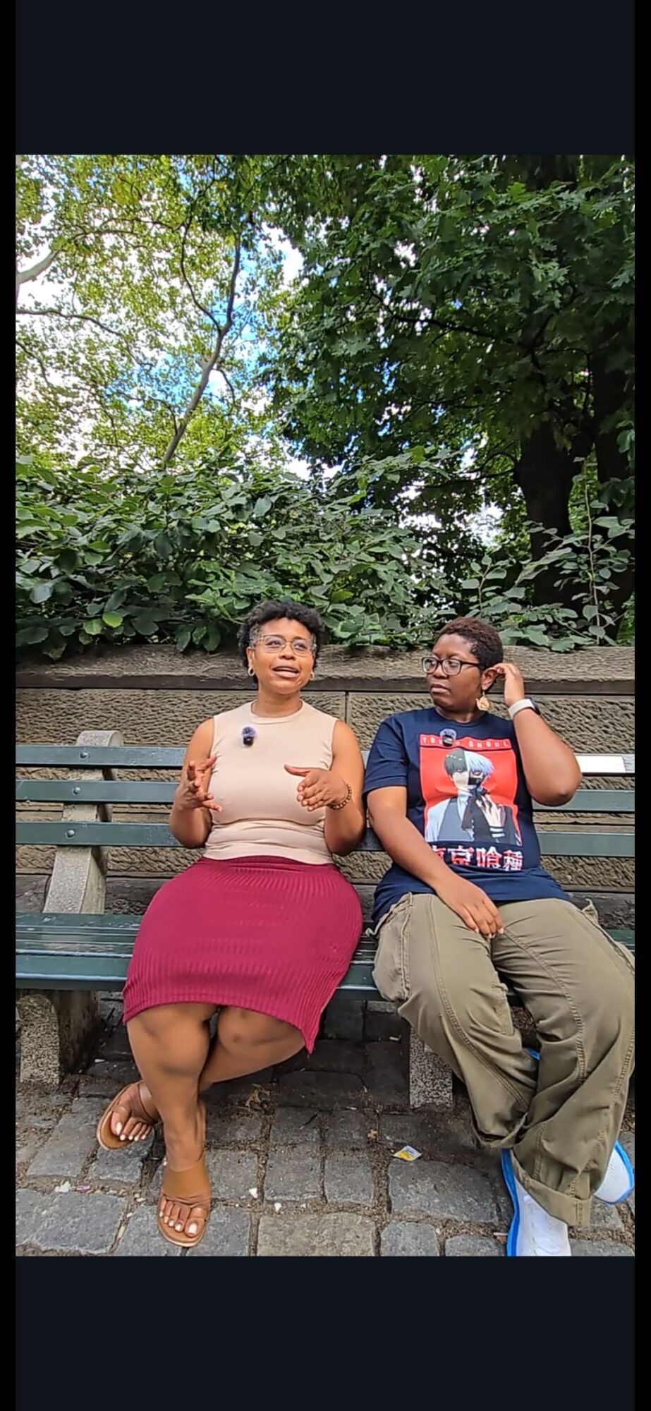 Two children sit on a park bench surrounded by green trees, one making a peace sign and the other adjusting glasses.