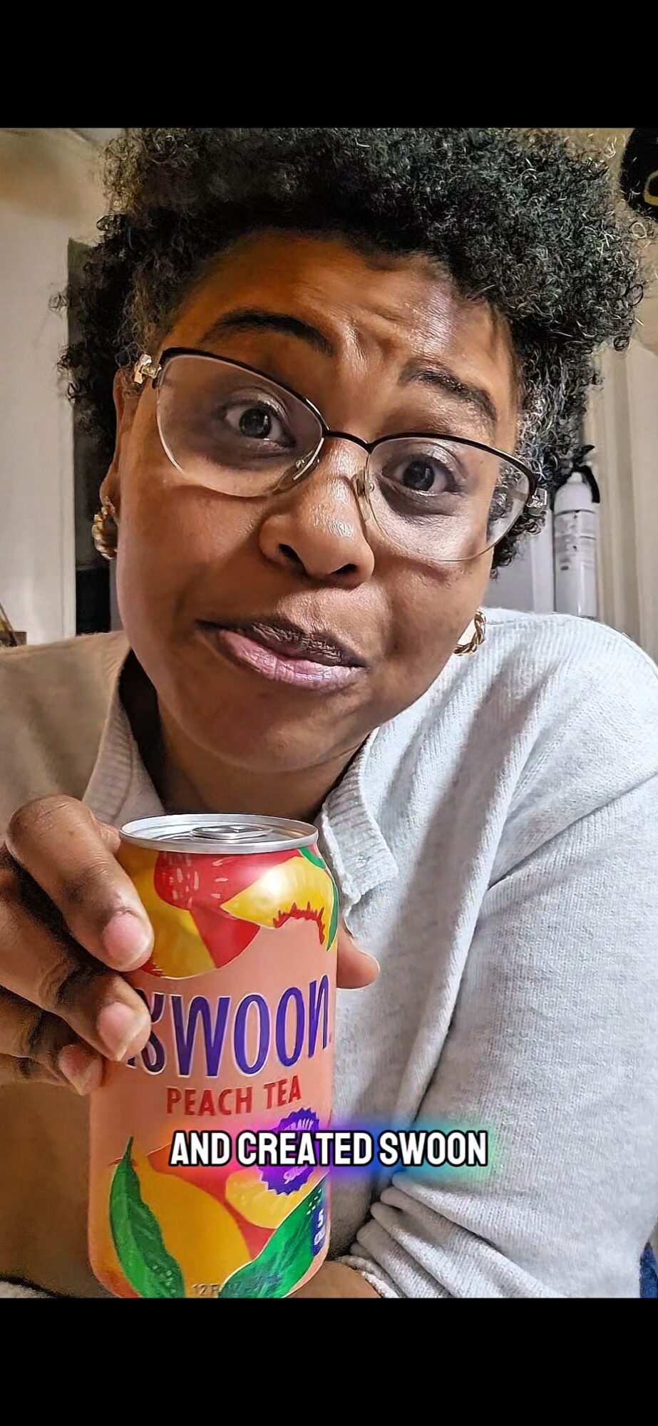 Person with glasses holding a can of peach tea, making a surprised expression, indoors.