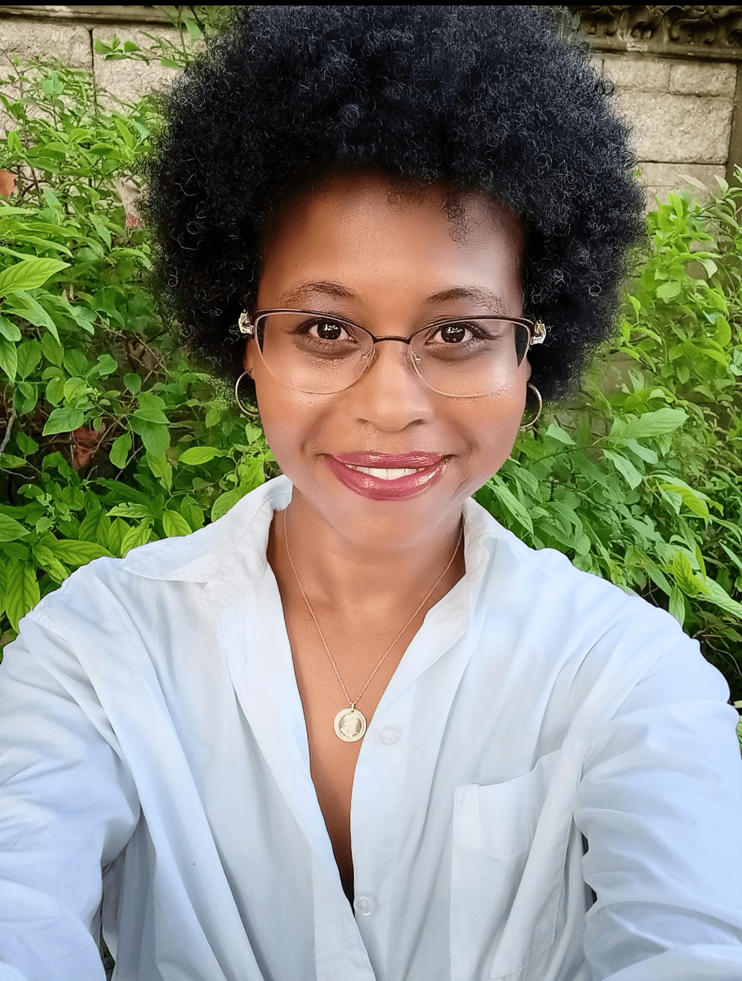 Woman with glasses and curly hair smiling outdoors, wearing a white shirt and a necklace, with green foliage background.