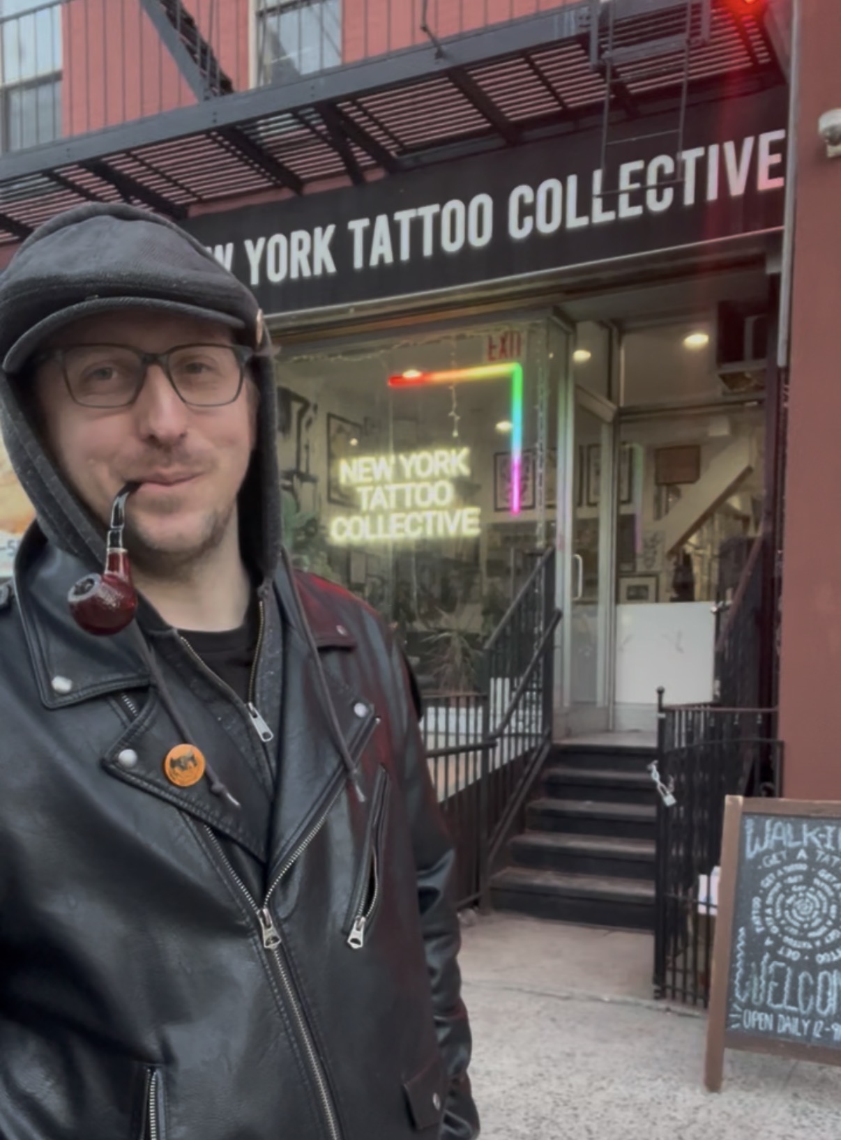 Man with glasses and a cap standing outside tattoo shop with neon sign, stairs, and chalkboard sign, daytime.