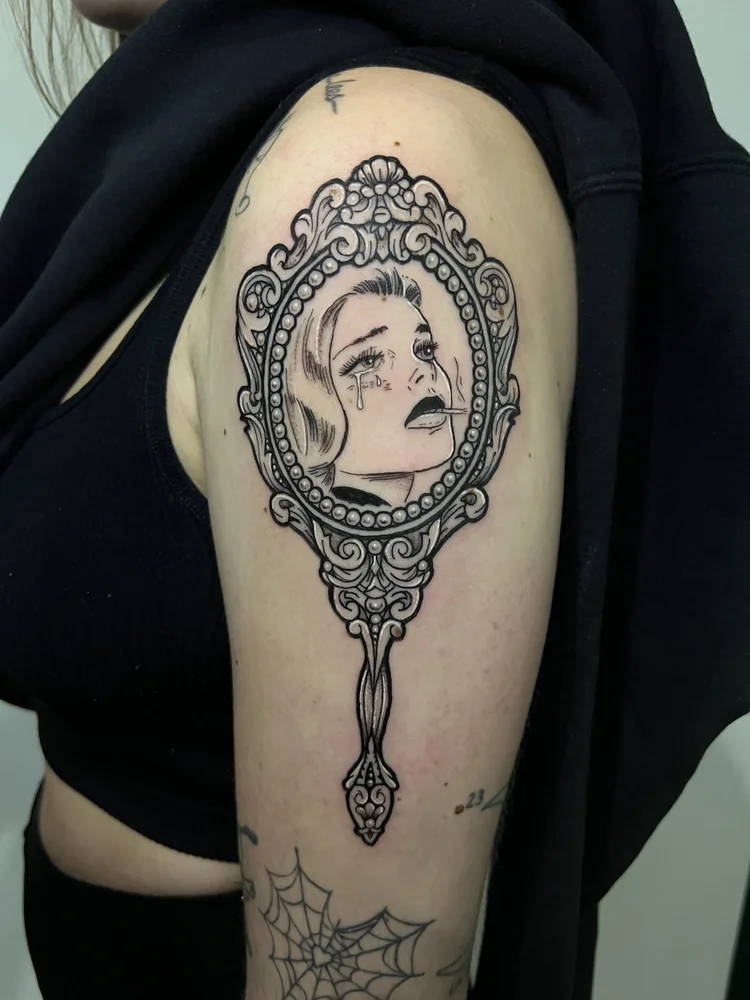 Tattoo of a woman's face in an ornate mirror frame on upper arm, with a spider web below.