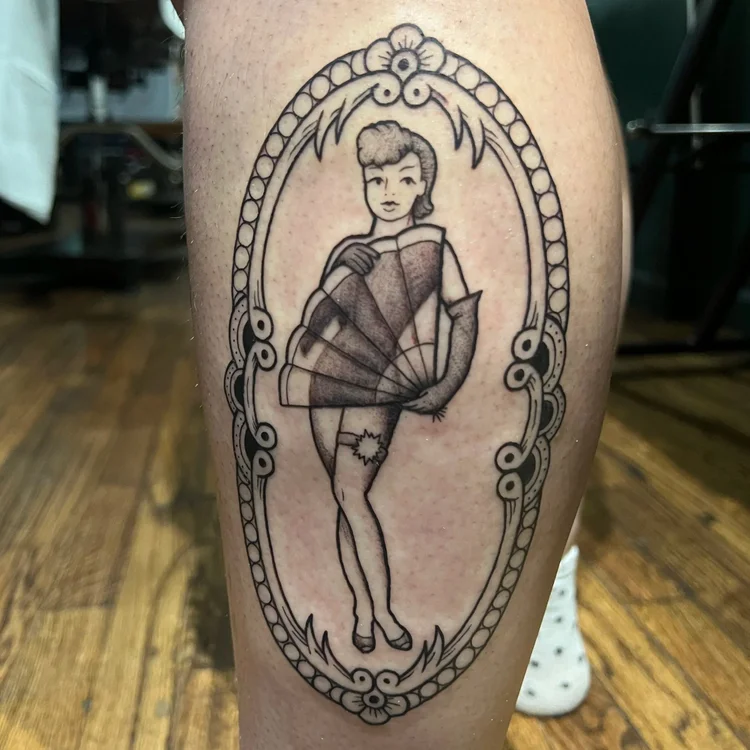 Tattoo of a woman holding a fan, framed by an ornate oval border, on a person's leg.