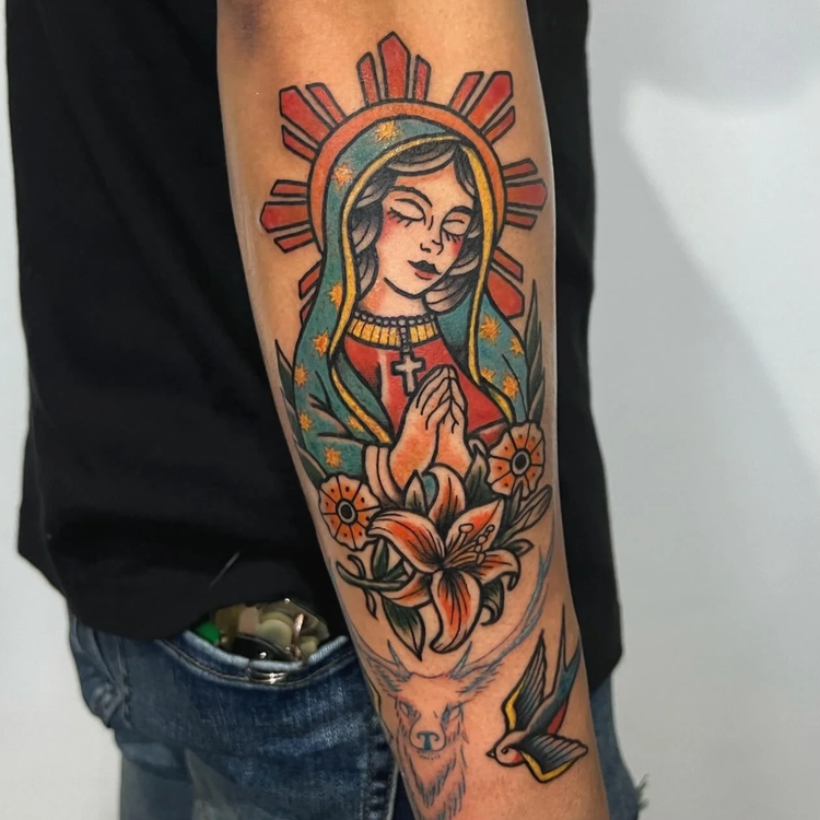 Tattoo of a woman with a halo, hands in prayer, surrounded by flowers and a bird on the arm.
