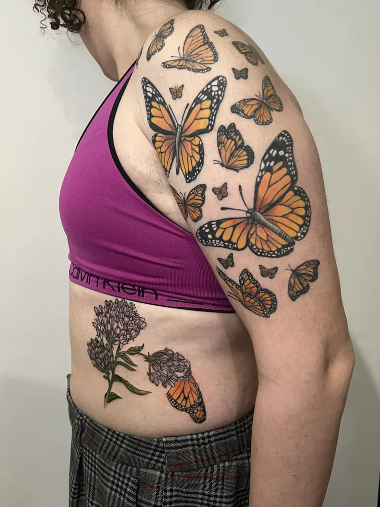 Person's upper arm and side with colorful butterfly tattoos, wearing a pink sports bra and plaid pants.