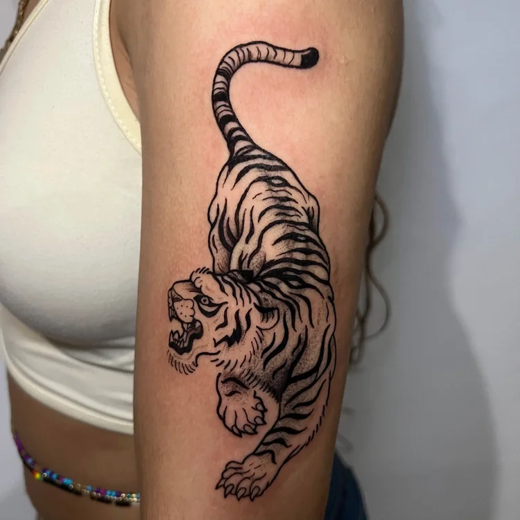 Black and gray tiger tattoo on upper arm, showing tiger in a crouching position with detailed stripes and facial features.