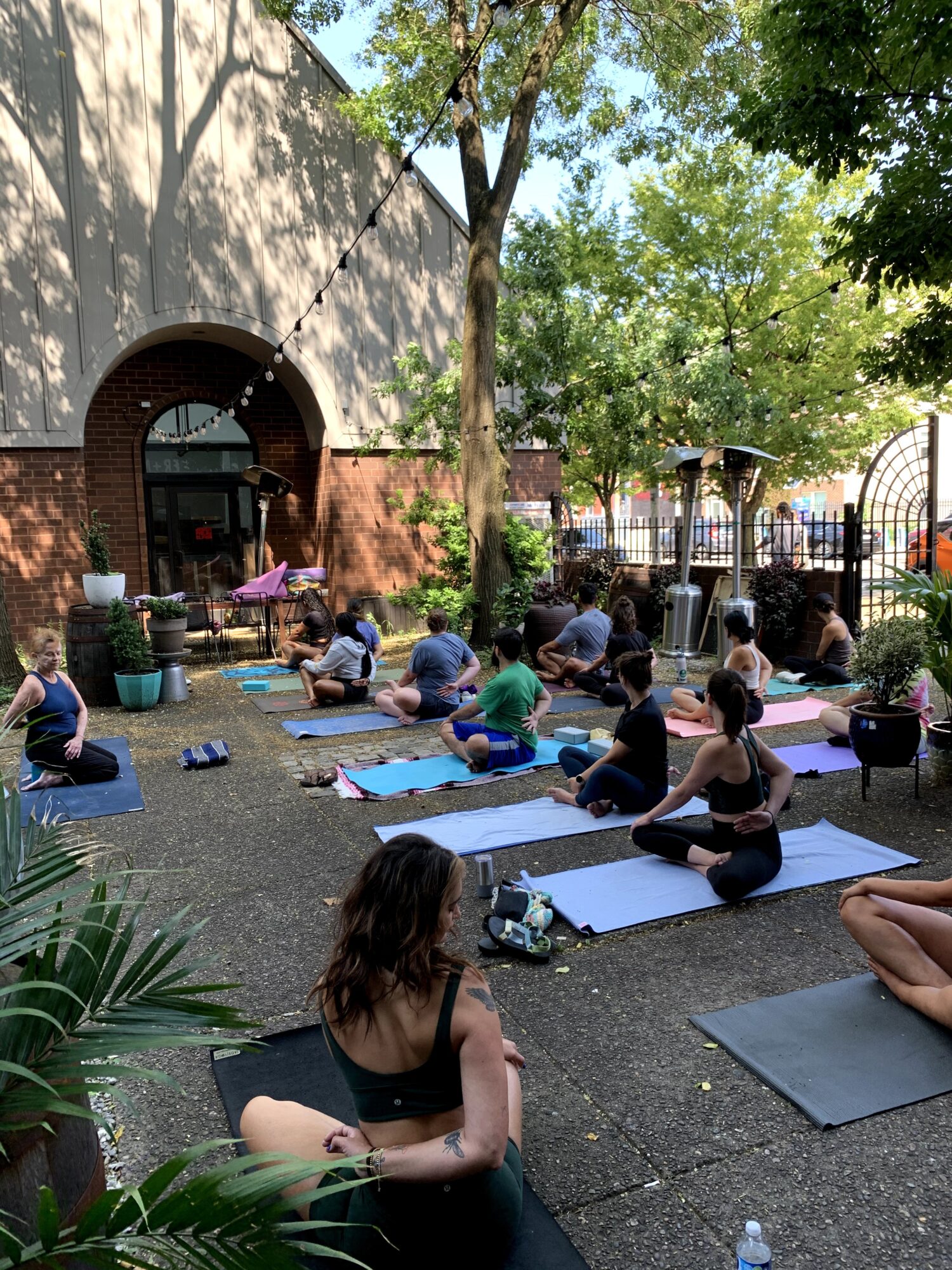 People practicing yoga outdoors on mats in a garden with trees and a building in the background.
