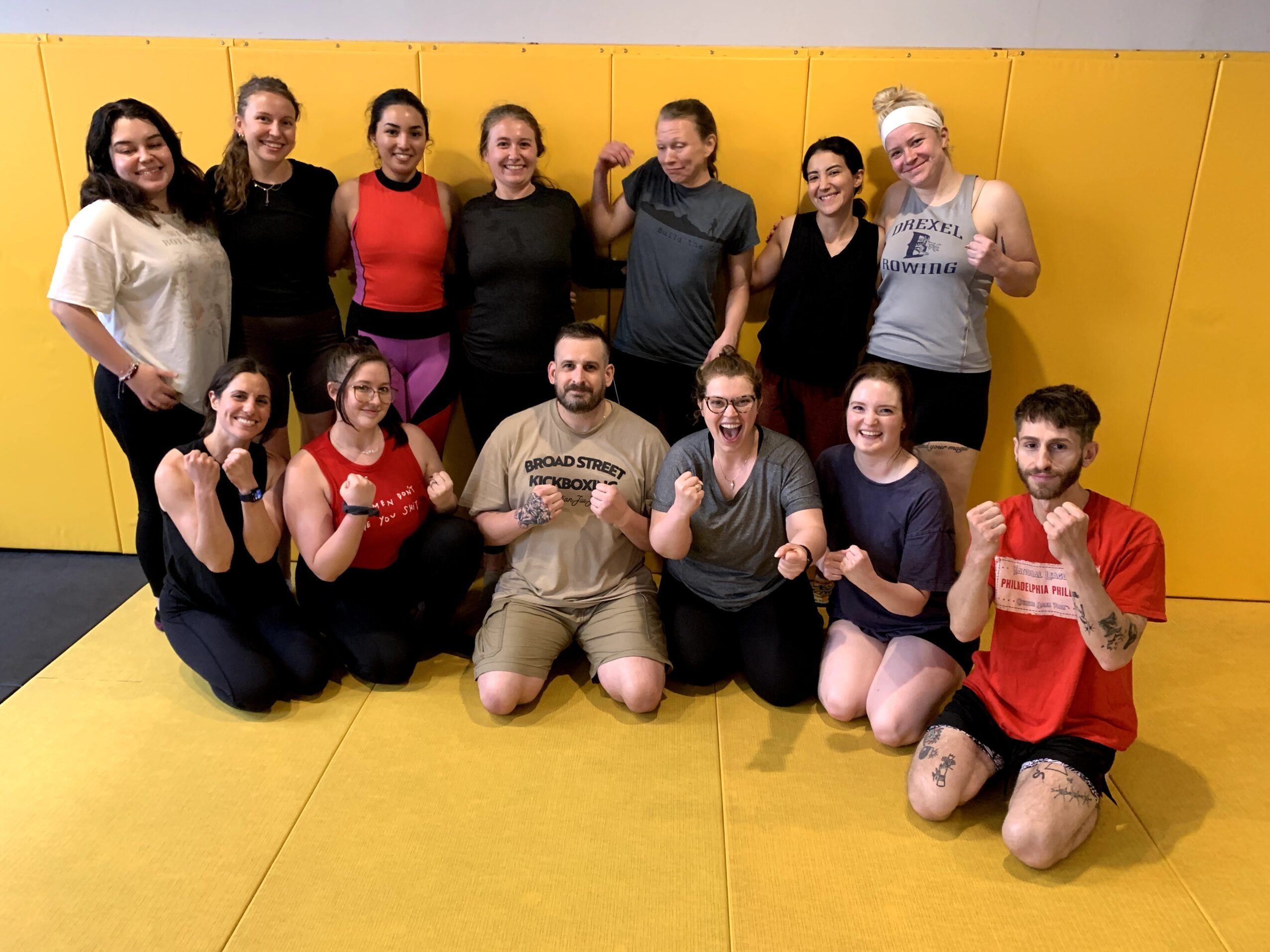 Group of 13 people in a gym, some showing fists, standing and sitting on yellow mats, smiling, with a yellow wall background.