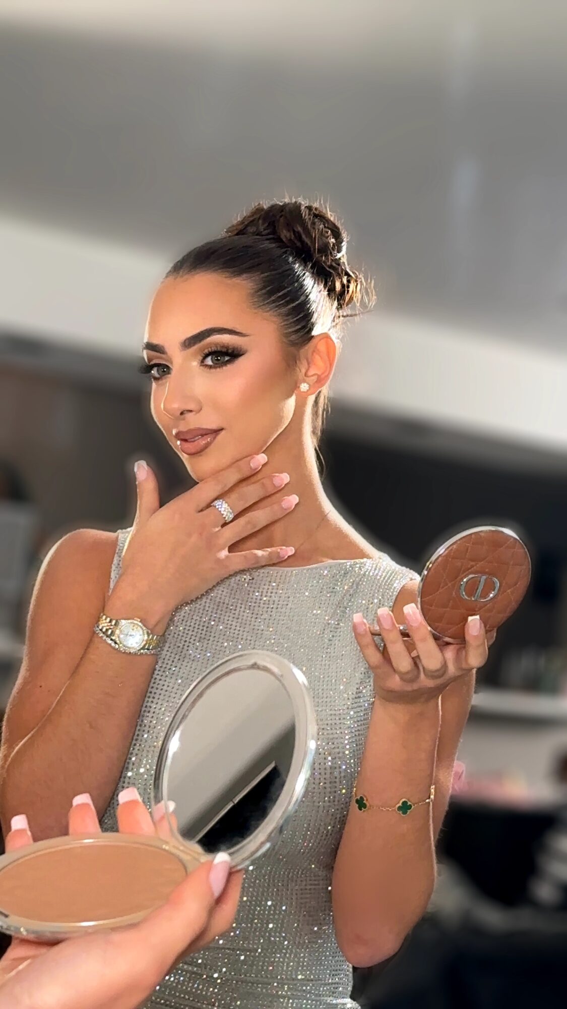 Woman with dark hair in a bun, makeup, and a sparkly dress, holding a compact mirror and touching her face.