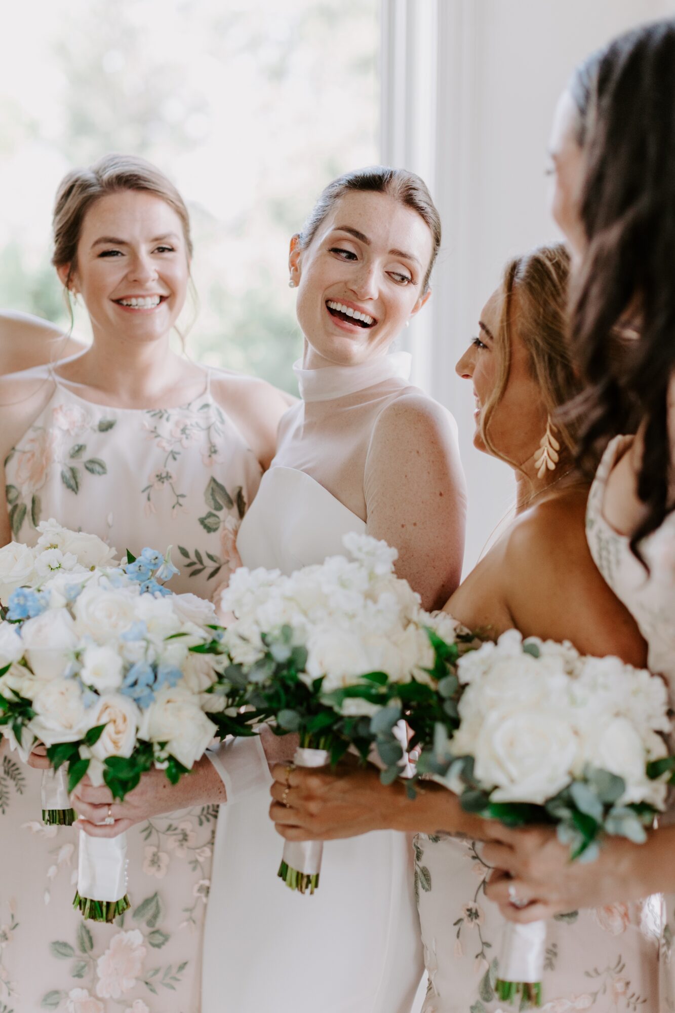 Four women in wedding dresses holding bouquets, smiling and talking indoors with a window in the background.