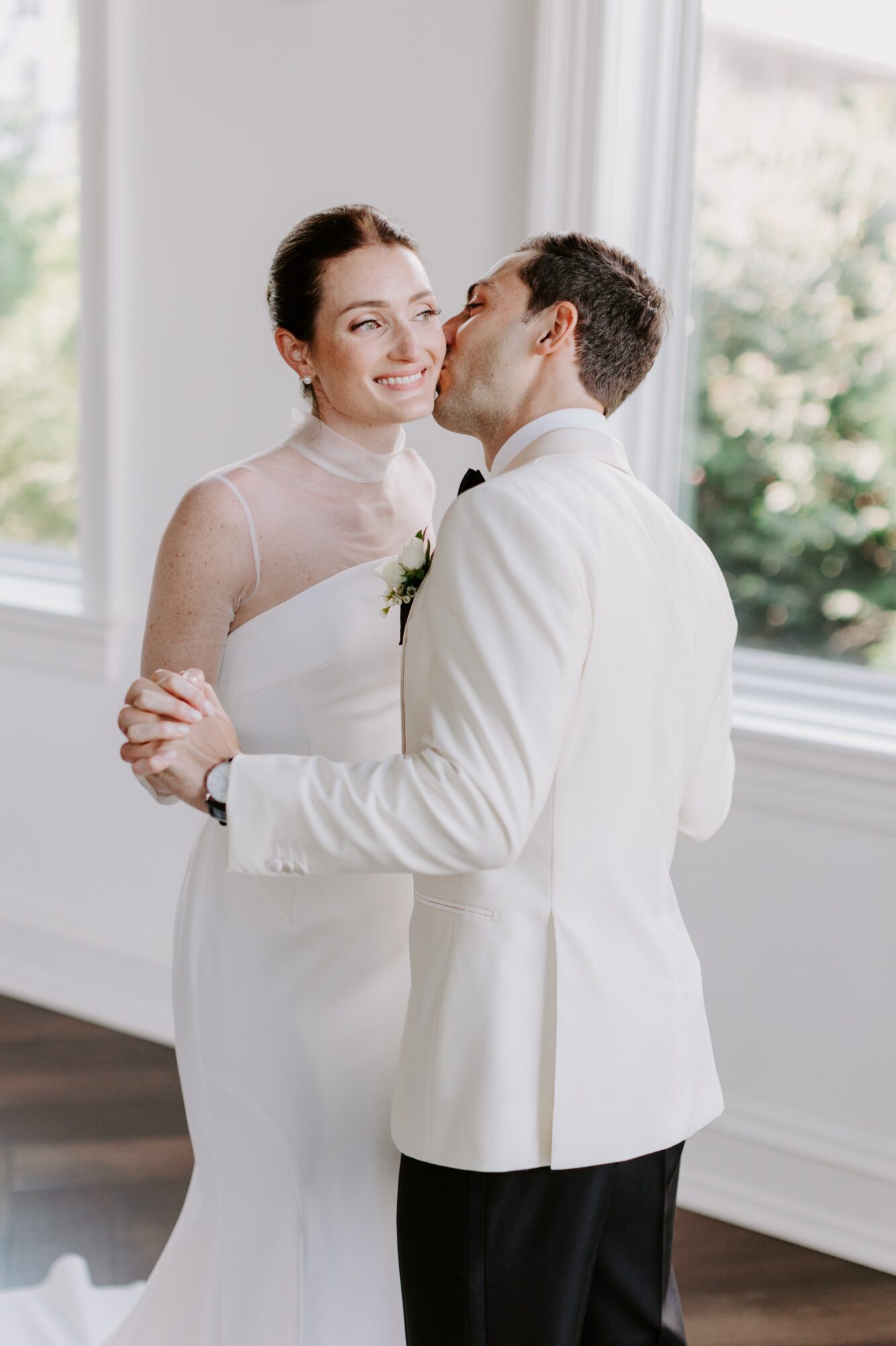 Bride and groom in wedding attire sharing a moment indoors with large windows and greenery outside, smiling and embracing.