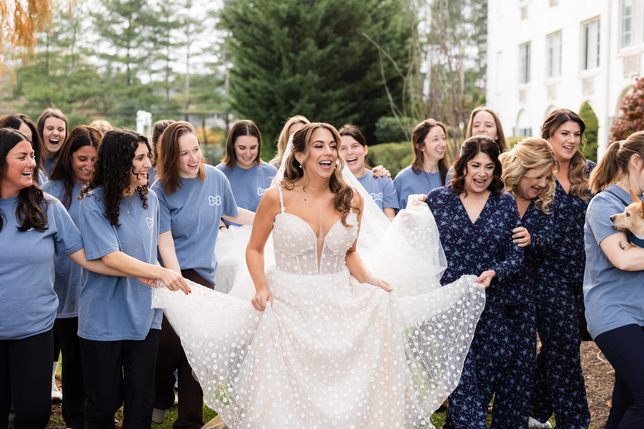 Group of women, some in matching blue shirts, gather outdoors around a smiling woman in a white dress holding her dress's sides.