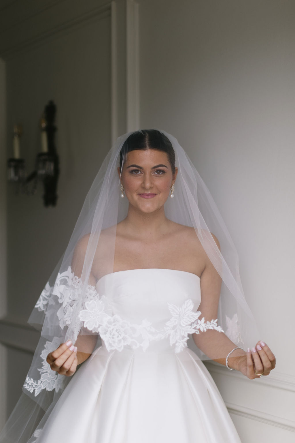Young woman in wedding dress with veil, smiling, standing indoors against a plain wall.