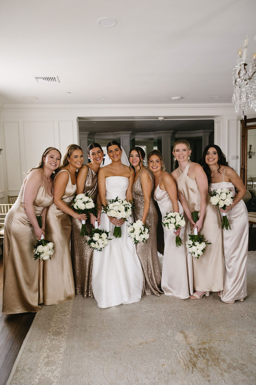 Group of seven women in elegant dresses, holding bouquets, standing indoors in front of a window and chandelier.