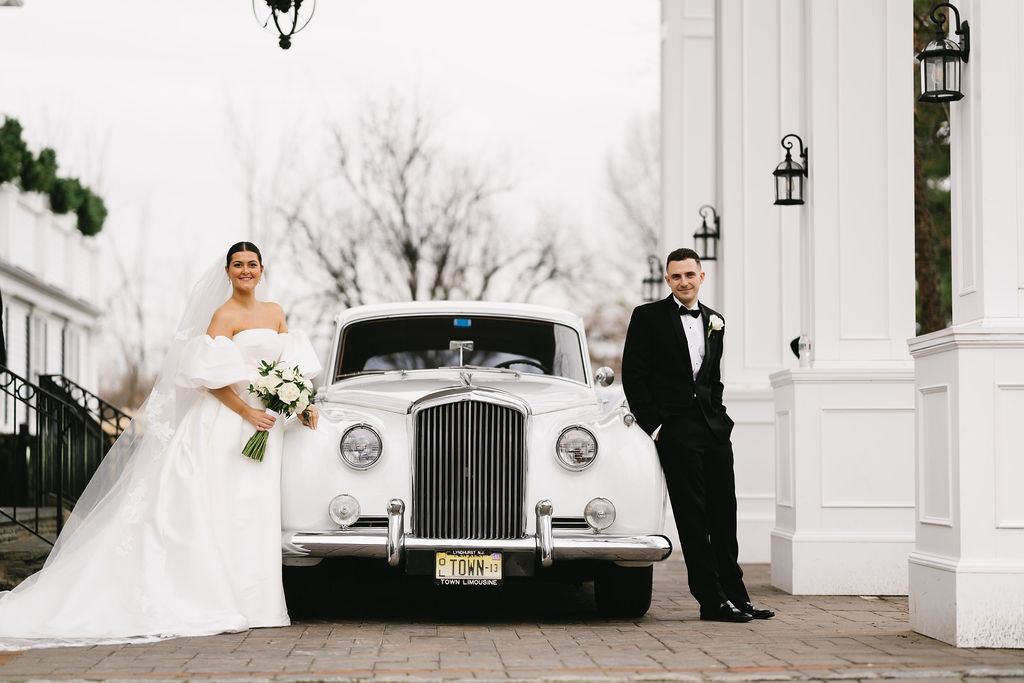 Bride and groom stand beside a vintage white car outdoors, with white columns and lanterns in the background.