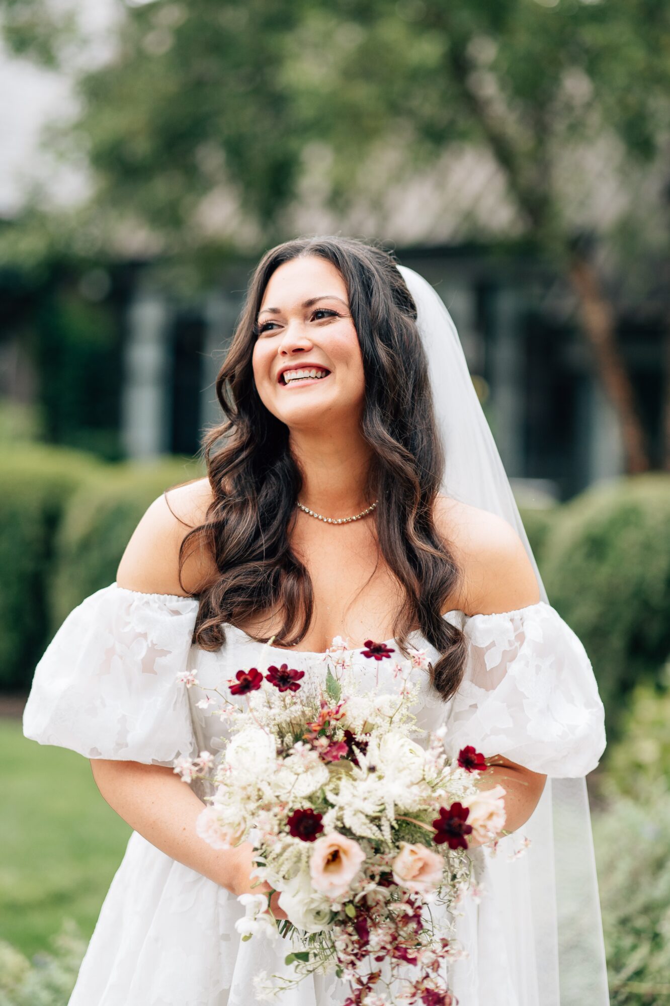 Smiling woman in wedding dress holding a bouquet outdoors, with trees and greenery in background.