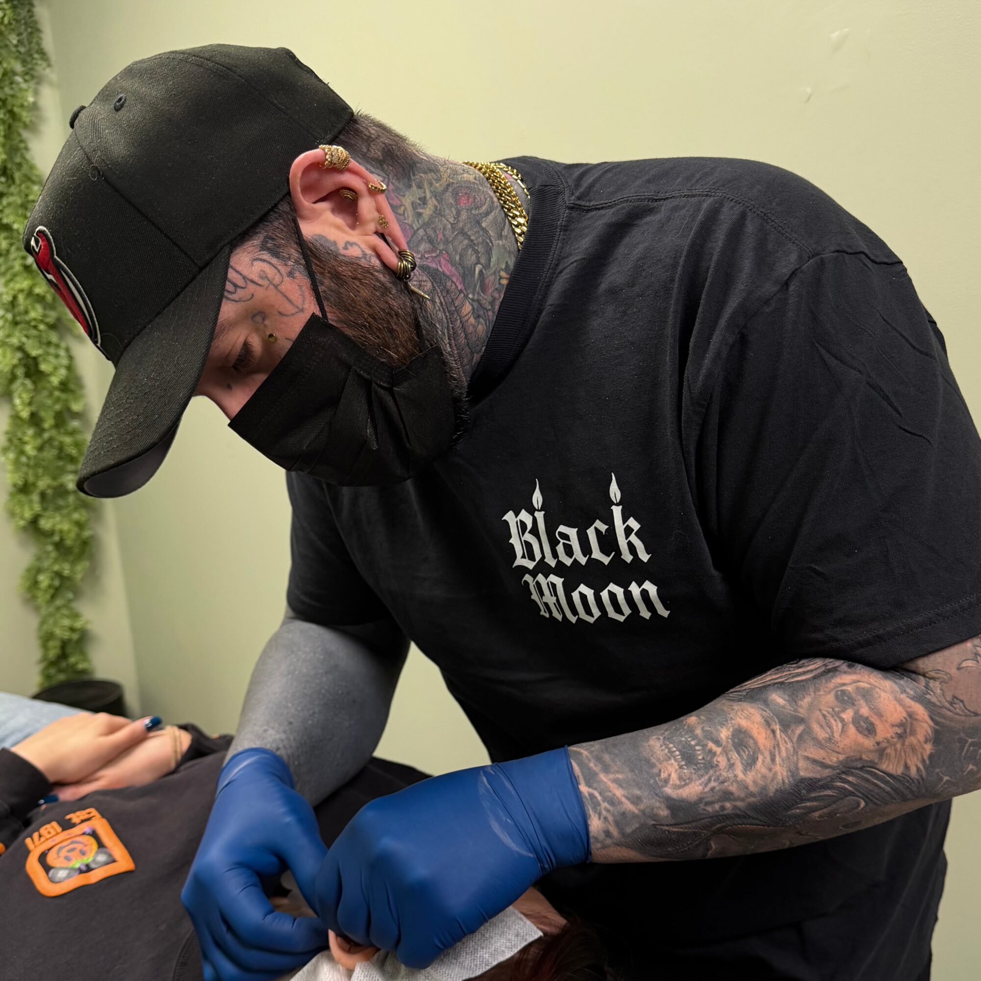 Tattooed man wearing a black cap, mask, and shirt with 'Black Moon' text, receiving a tattoo with blue gloves.