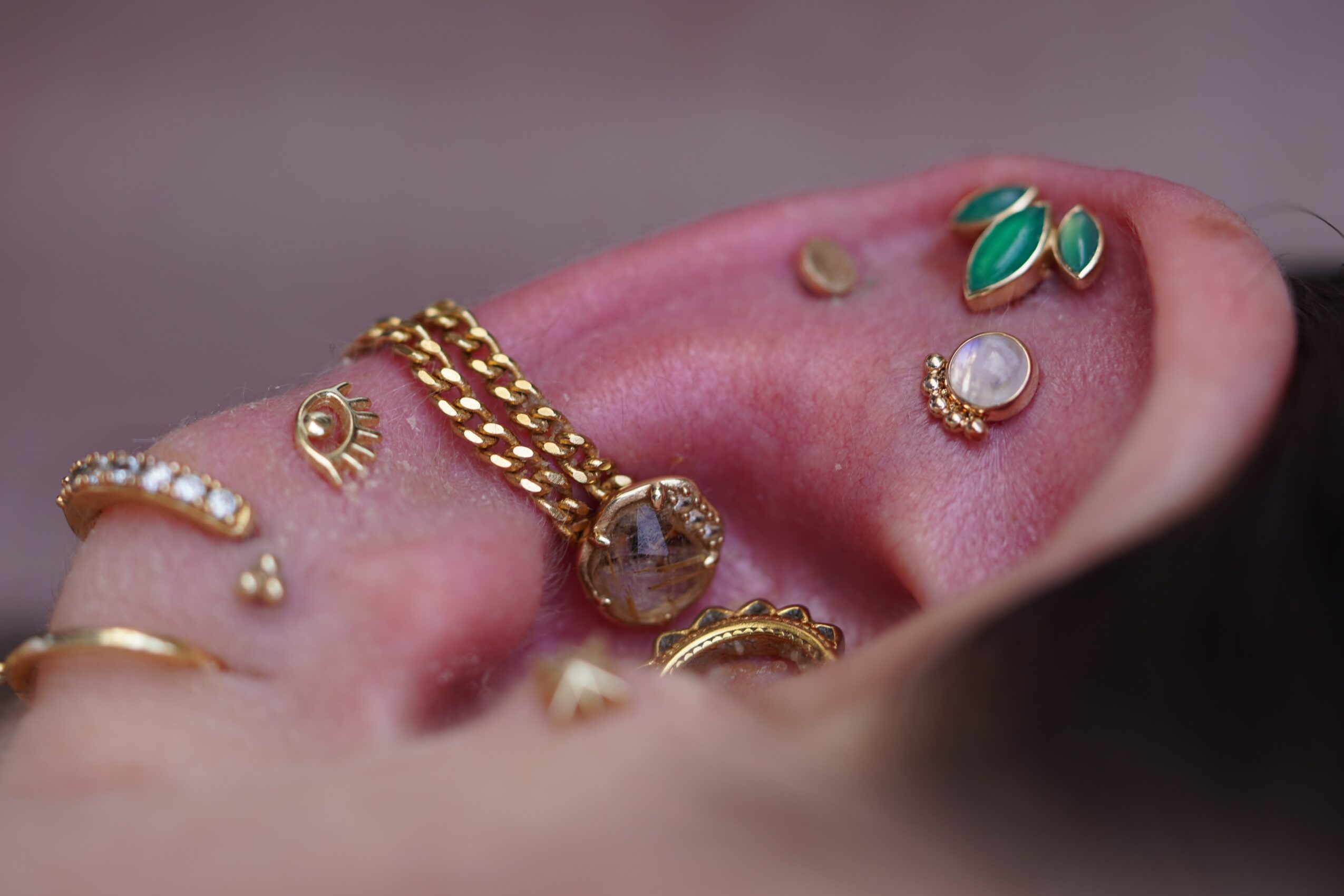 Close-up of a person's ear with multiple gold and gemstone earrings and jewelry, including a chain and decorative studs.