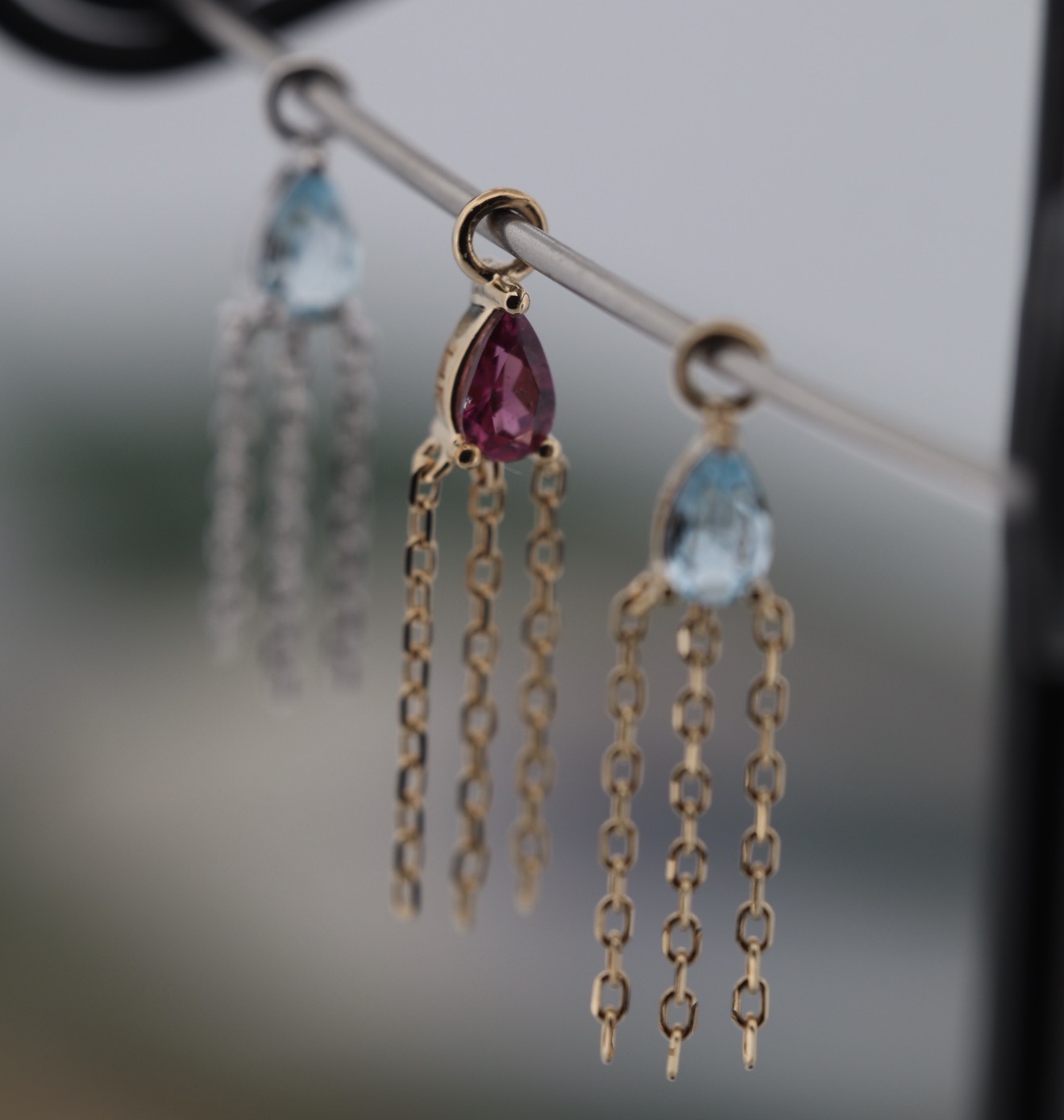 Three pairs of dangling earrings with colorful gemstones and chain tassels hanging from a horizontal bar.
