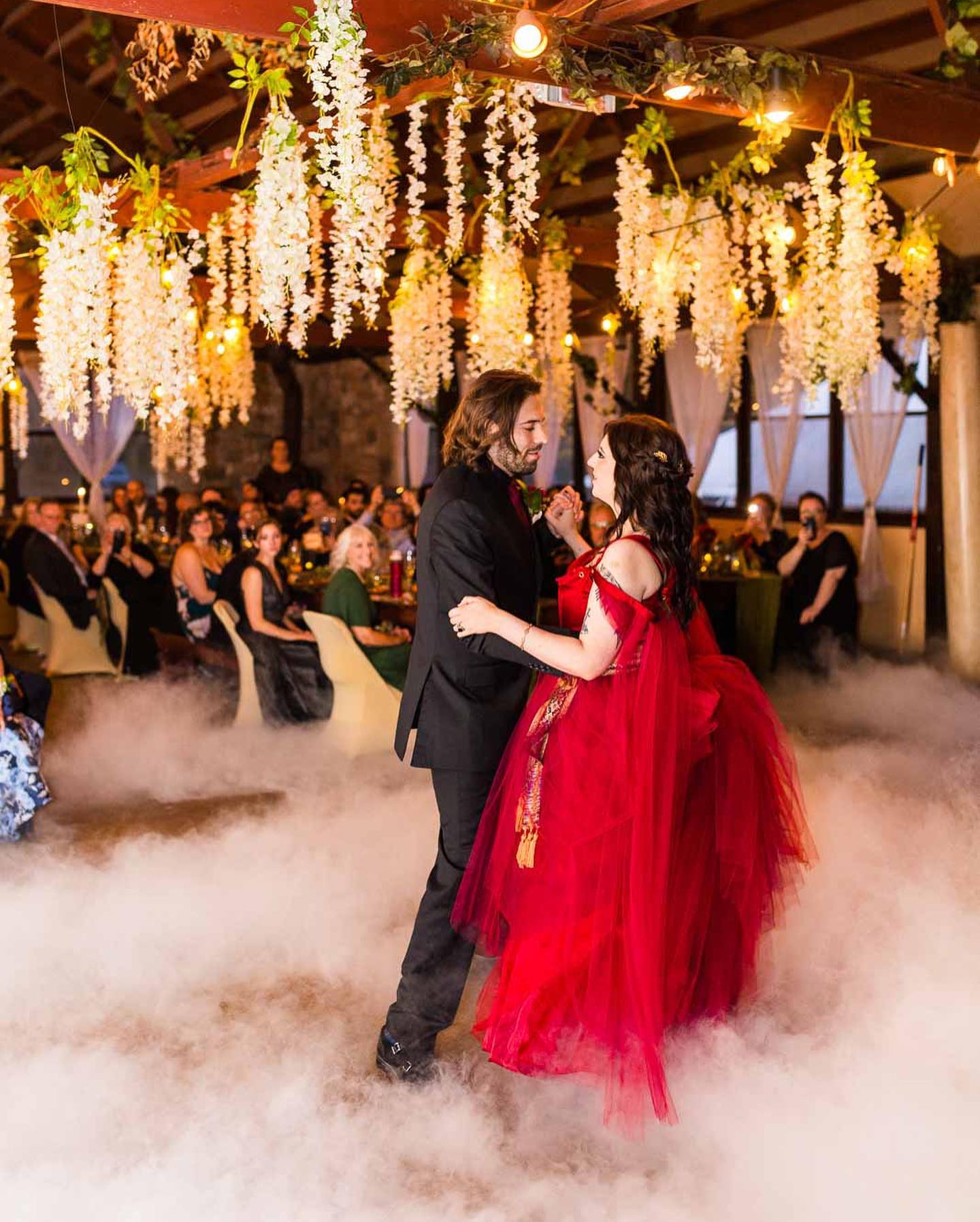 Couple dancing in formal attire under hanging white flowers and string lights, surrounded by seated guests, with fog on the floor.