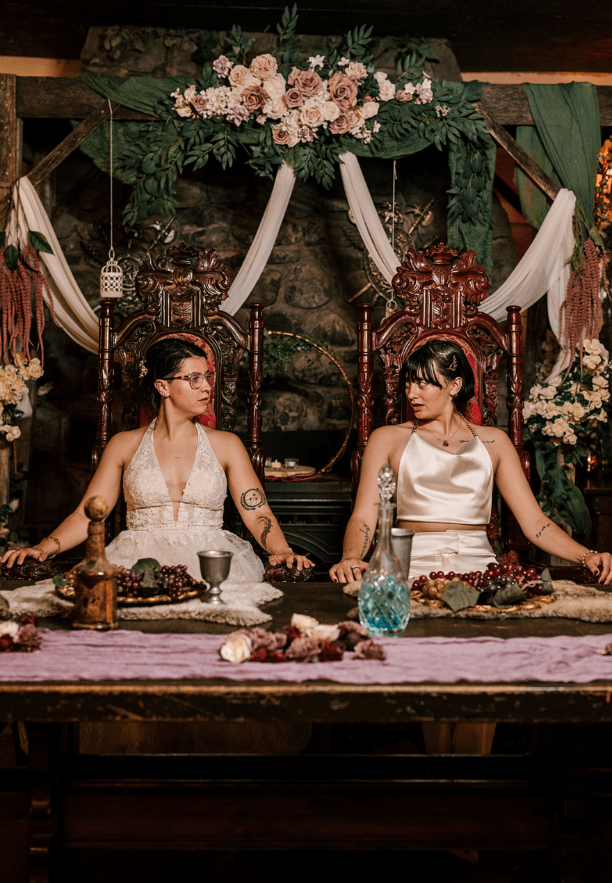 Two women sit at a decorated table with floral arrangements and ornate chairs, wearing white dresses, in a festive setting.