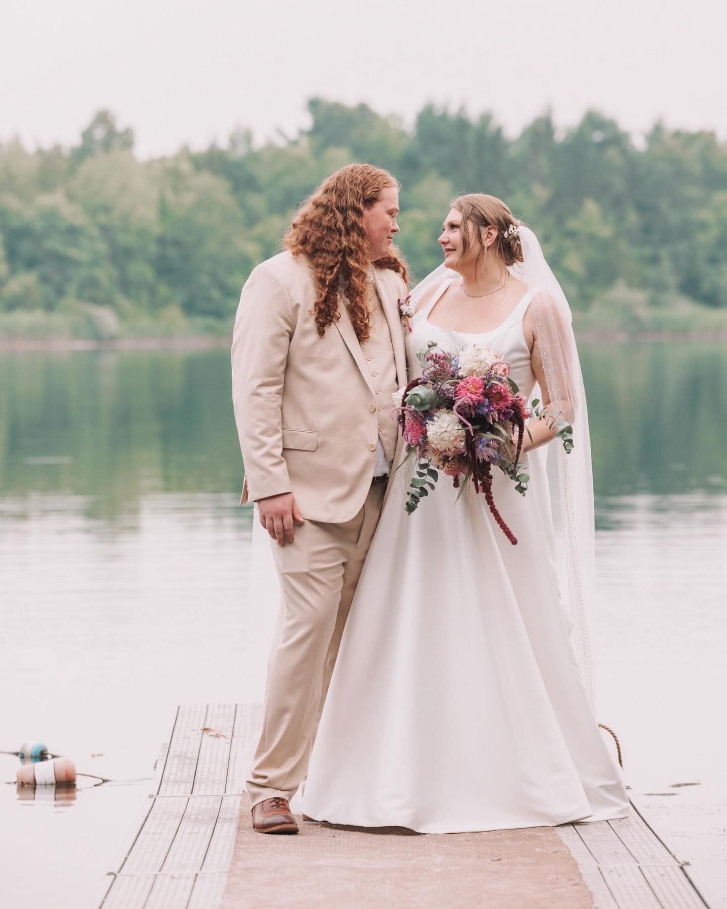 Bride and groom standing on a dock by a lake, holding hands, with trees in the background.