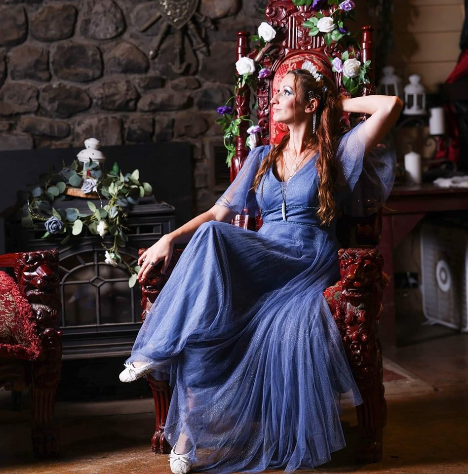 Woman in a blue dress sitting on a decorated chair with flowers and a stone wall background.
