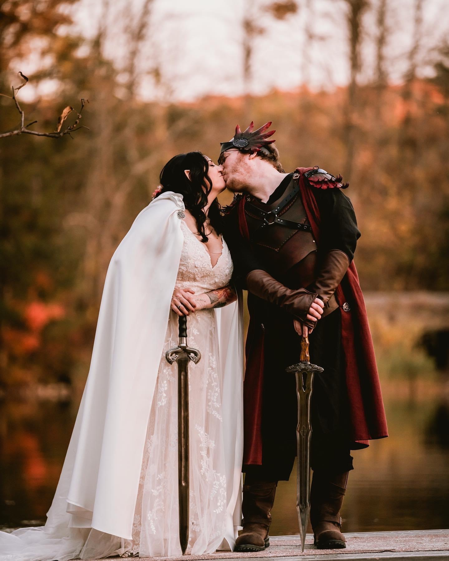 Two people in fantasy costumes kiss outdoors, holding swords, with a blurred autumn background.