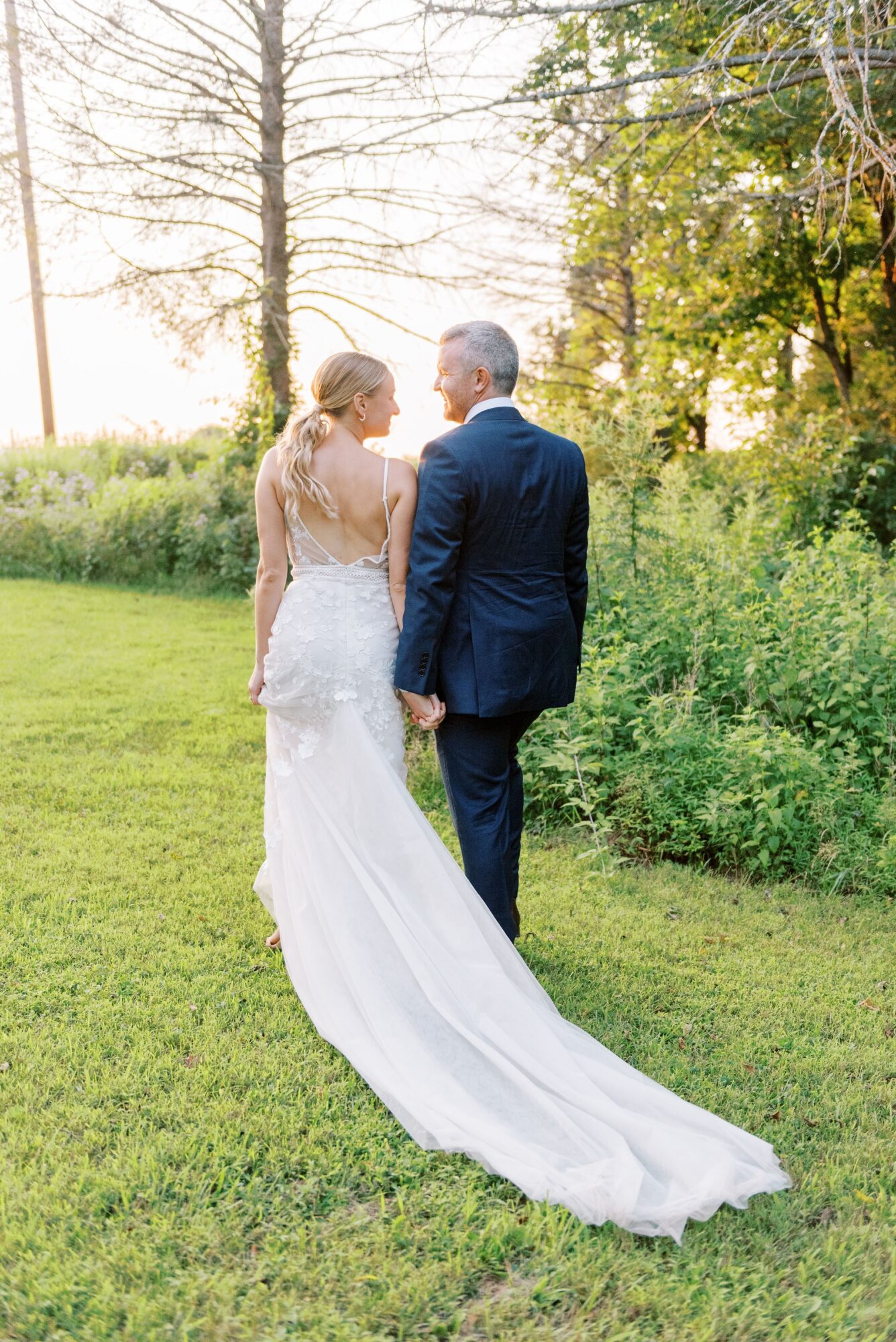 Bride and groom holding hands outdoors, facing away, with trees and grass in the background.